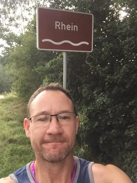 A man with glasses and a beard taking a selfie next to a sign that reads "Rhein" next to a river, with trees and greenery in the background.