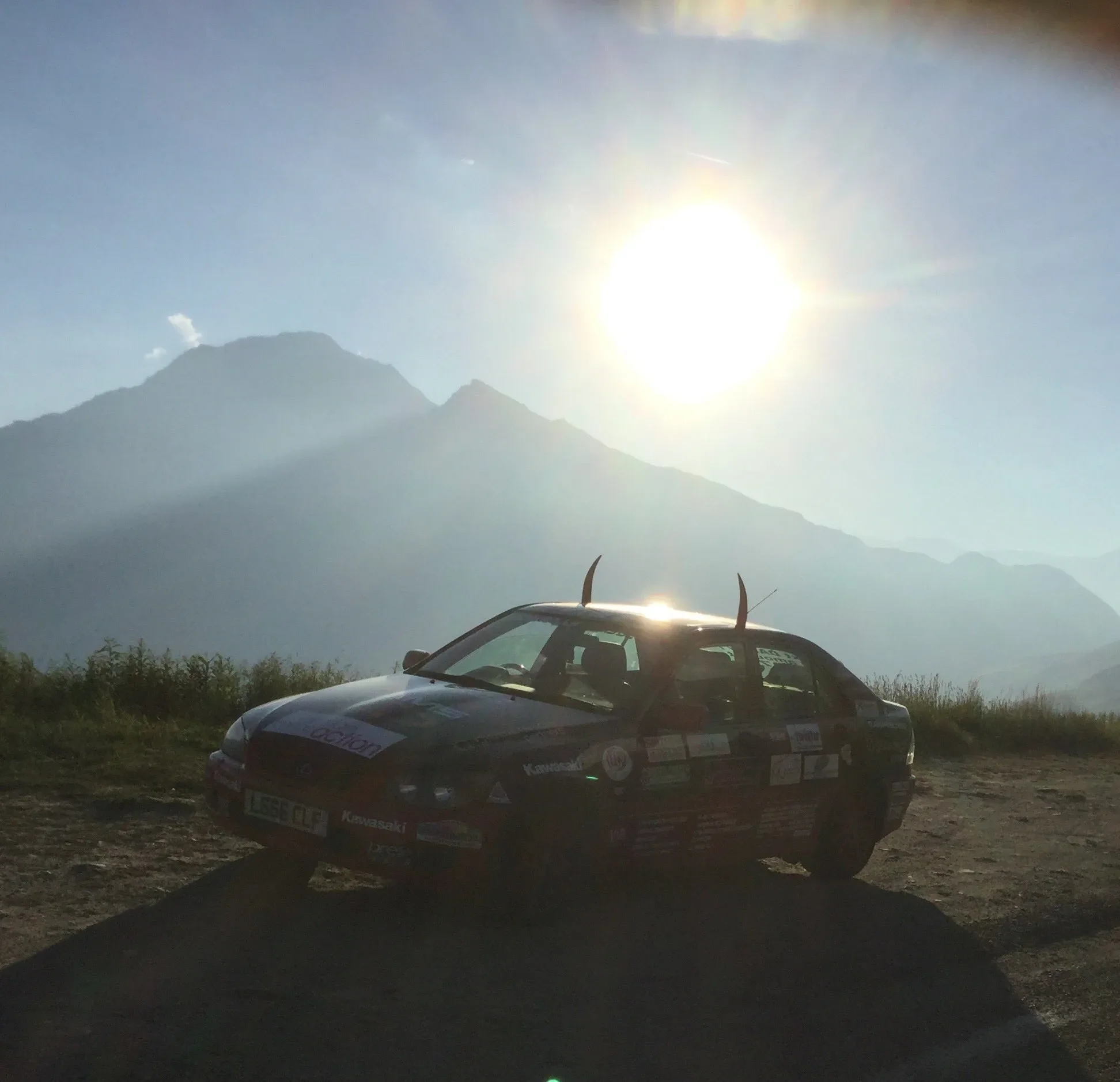 A rally car with decals parked on dirt ground with mountains and a bright sun in the background.