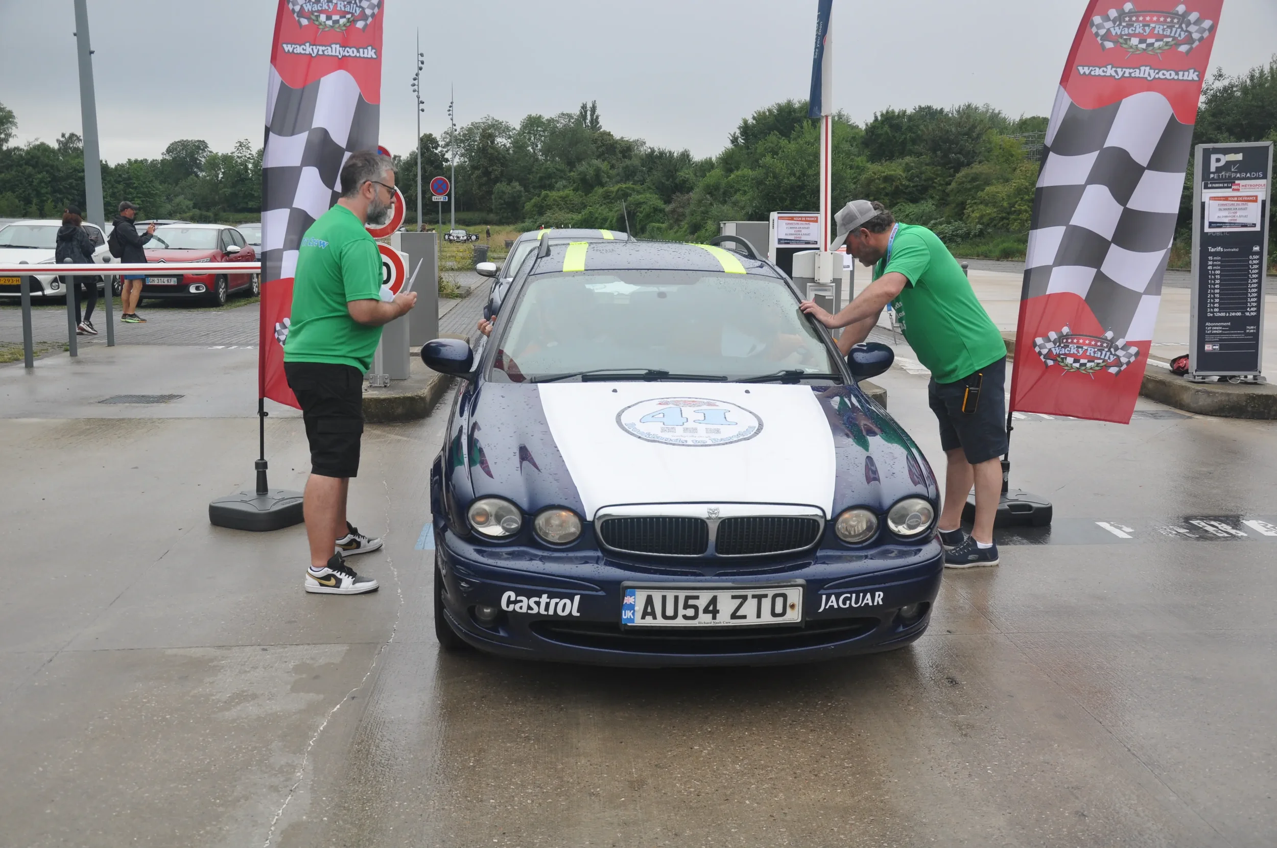 A classic blue Jaguar race car with a white hood, numbered 41, parked at a rally event with two men in green shirts working on it. Two red checkered flags with 'Wacky Rally' branding are on either side of the car, and other cars and spectators are in