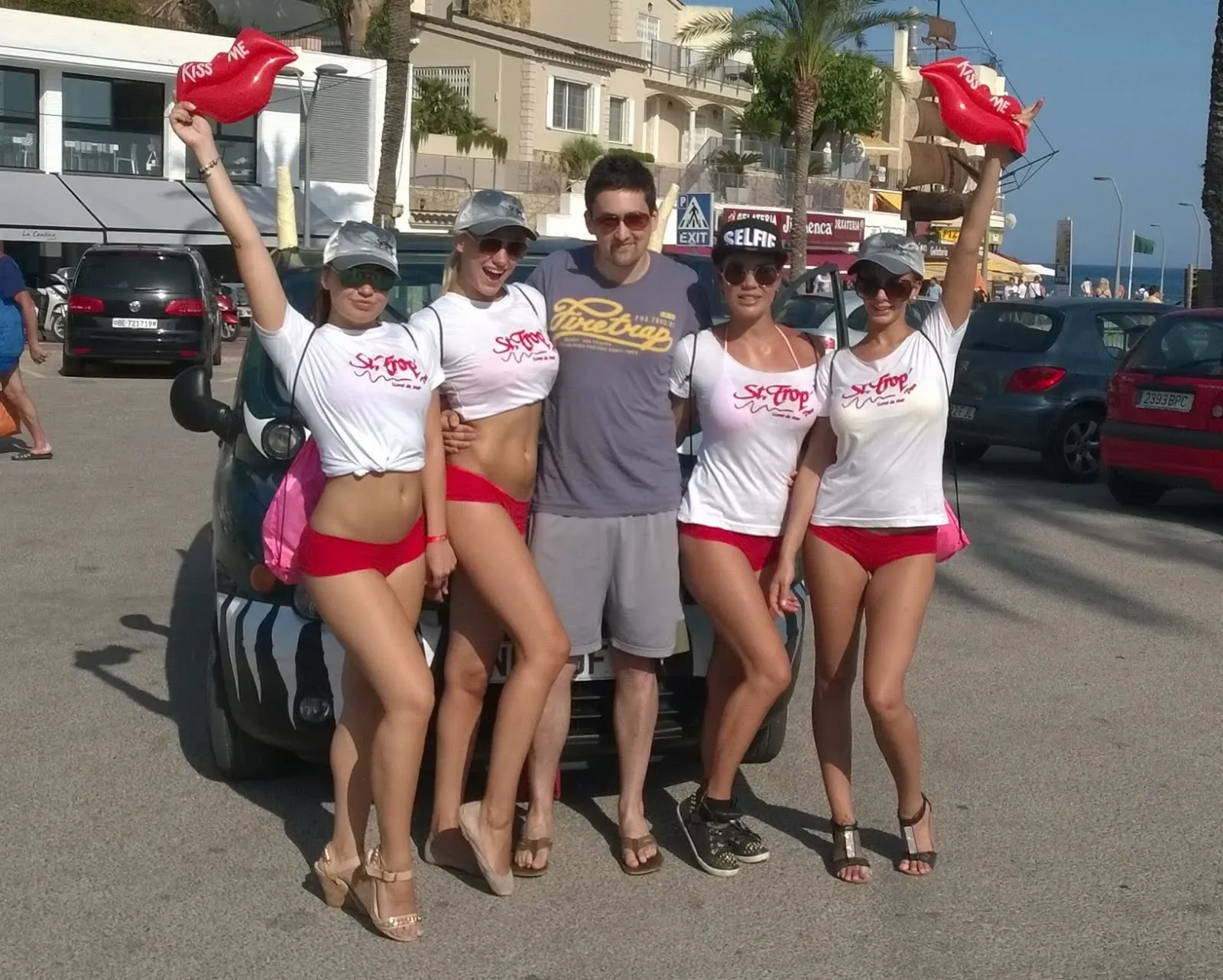 Four women in white T-shirts and red shorts posing with a man in gray shorts and T-shirt on a beachside street. The women are holding red inflatable lips with 'Kiss Me' written on them. They are wearing hats, sunglasses, and sandals, and are standing
