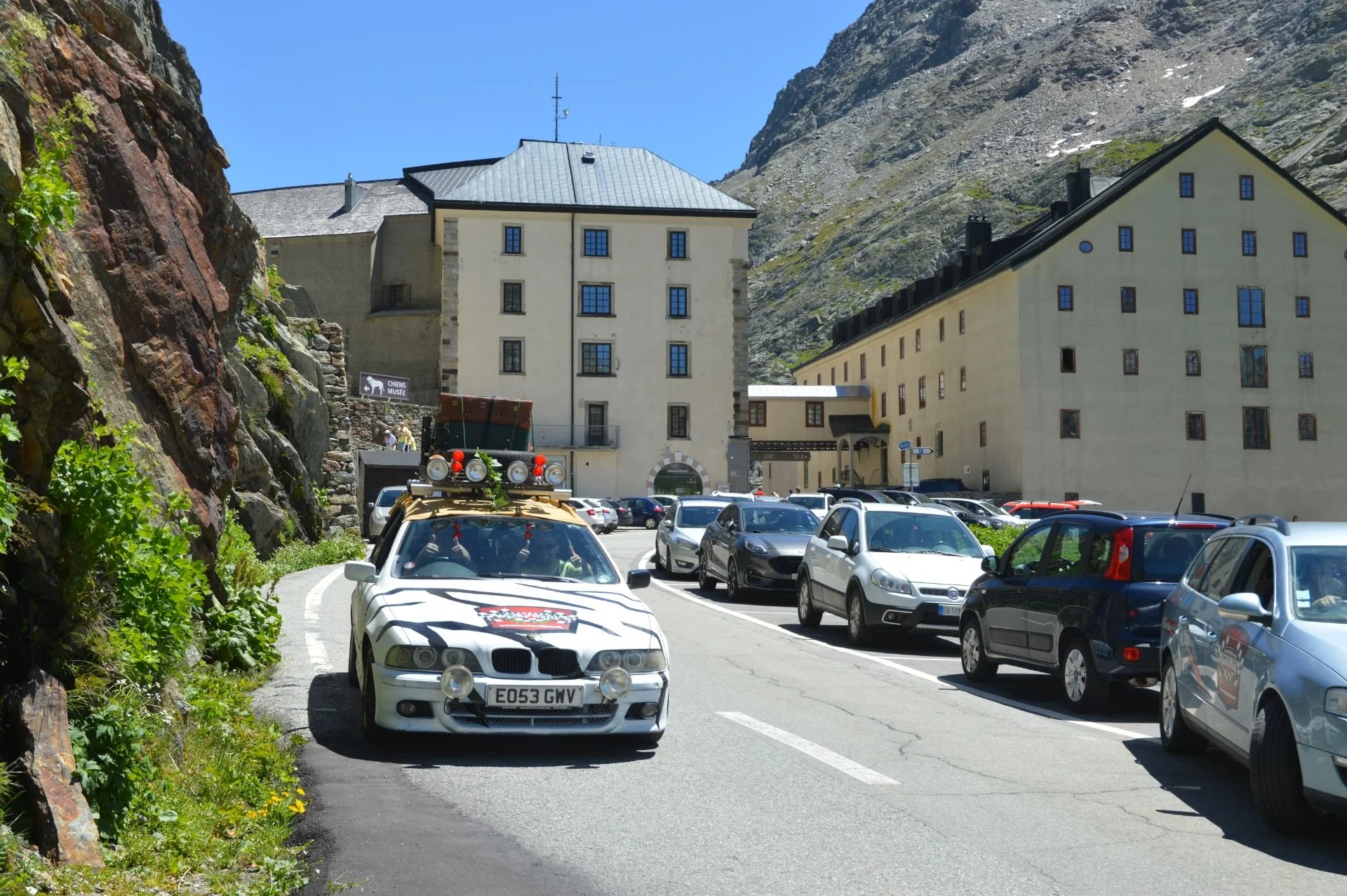 A parking lot near a mountain with several cars parked. A distinctive white car with a roof rack and mounted lights in the foreground. Large buildings with multiple windows and a hillside in the background.