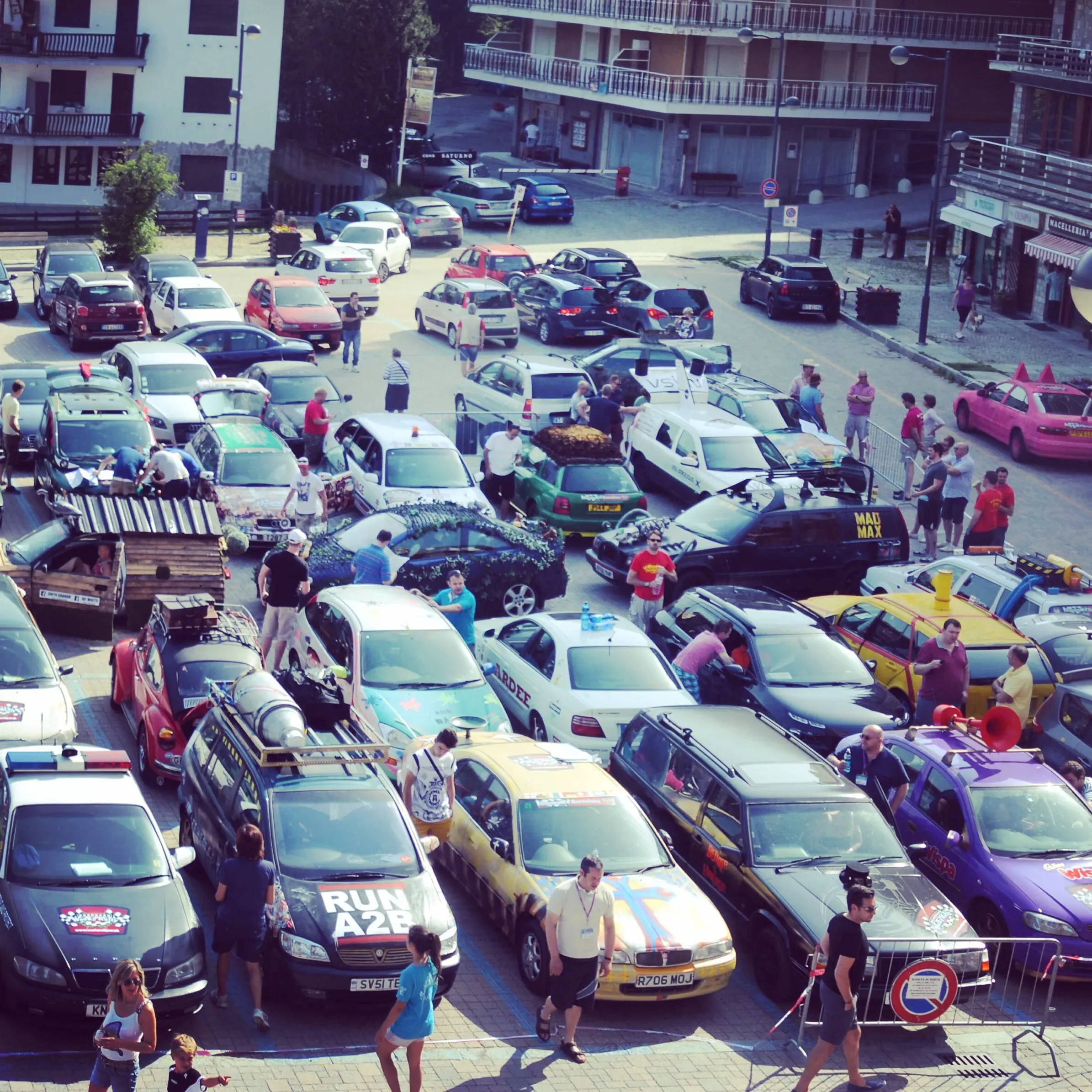 A crowded parking lot filled with various cars and people walking around, some interacting with the vehicles and others standing or talking, in an urban area with buildings and shops nearby.
