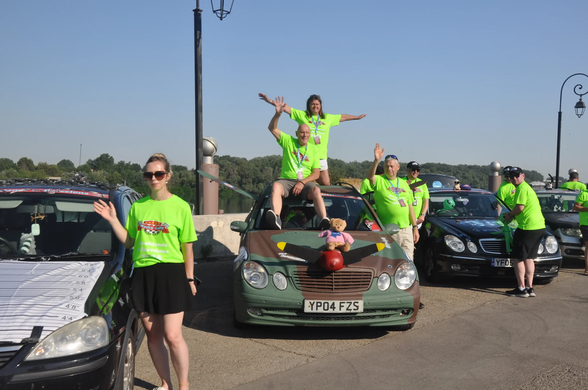Group of people wearing bright green shirts at a car rally, with some giving waves and others standing beside or on decorated cars near a body of water.