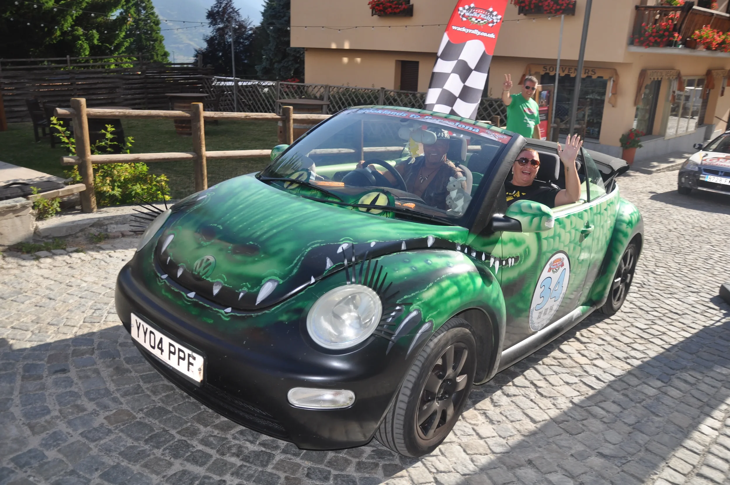 A green Volkswagen Beetle with a crocodile face and teeth painted on the front, parked on a cobblestone street. Inside, two women are smiling and waving, with one woman wearing sunglasses. In the background, a man in green is standing and waving near