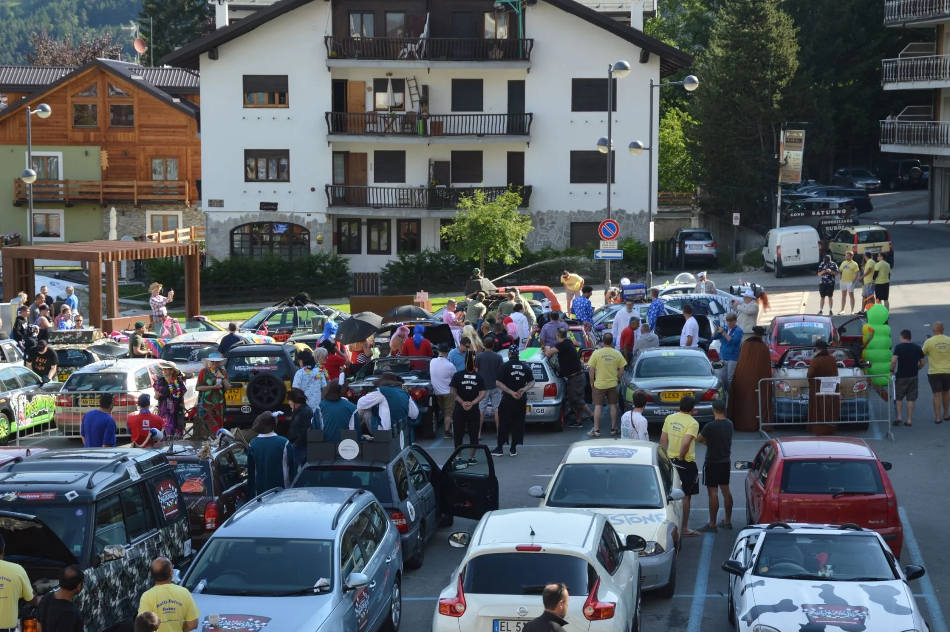 Crowd gathered around a lineup of cars in a parking lot during a daytime event in a mountain town, with houses and trees in the background.