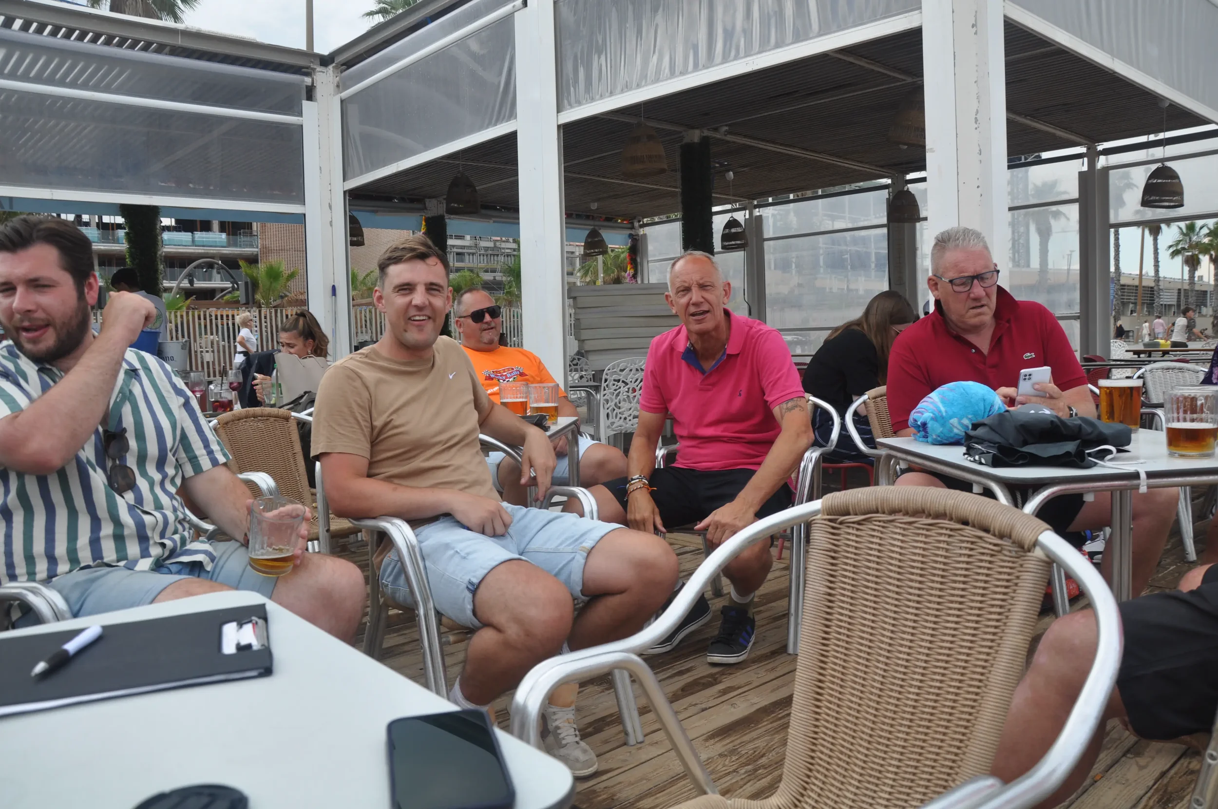 Group of five men sitting at a table on a covered outdoor patio, some holding drinks, with one looking at his phone. People in the background, some standing, with palm trees visible outside.
