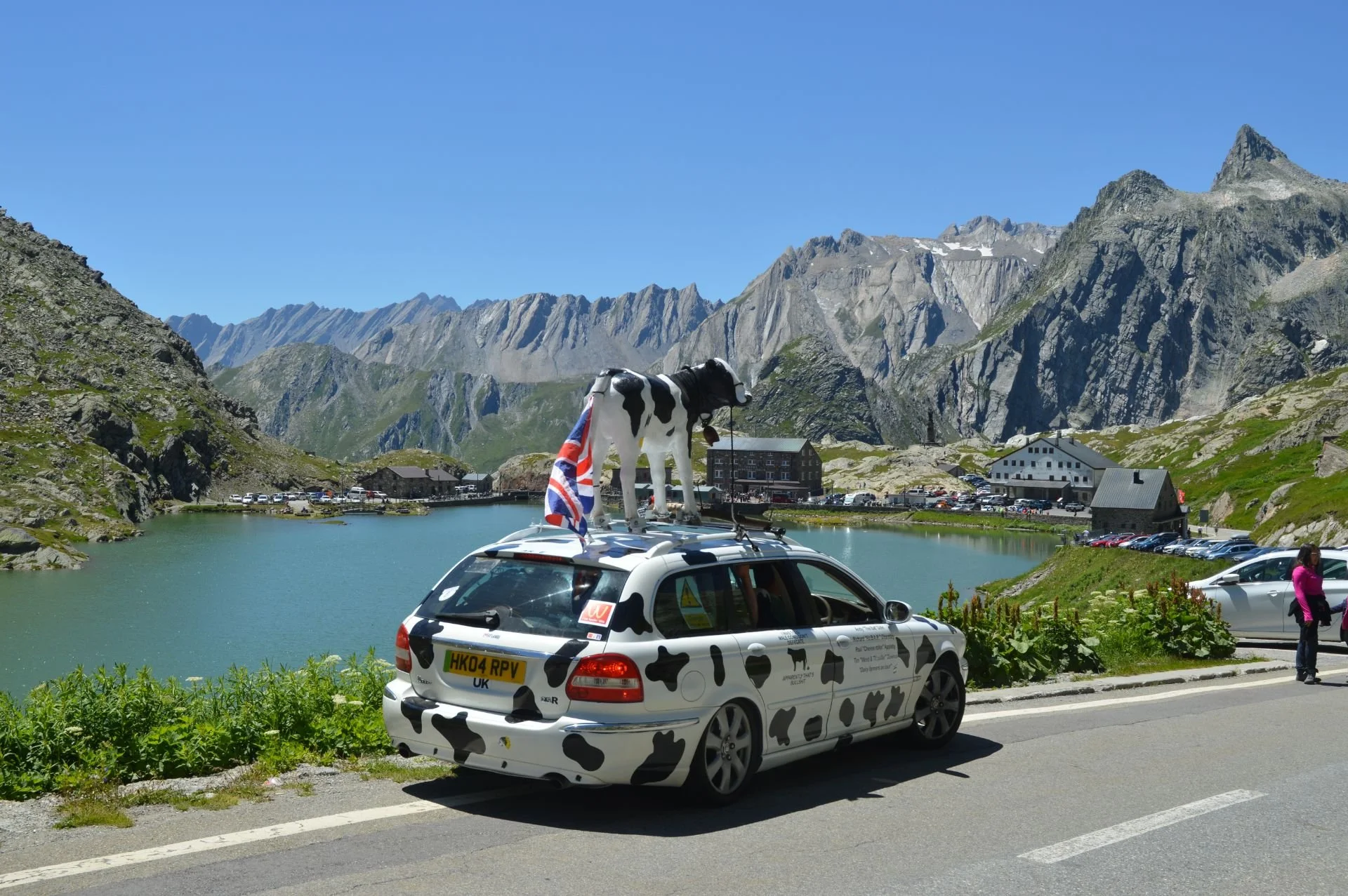 A car with cow print paint parked near a mountain lake, with a plastic cow on the roof and a Union Jack flag. The scene has mountainous terrain, some buildings, and a parking lot under a clear blue sky.