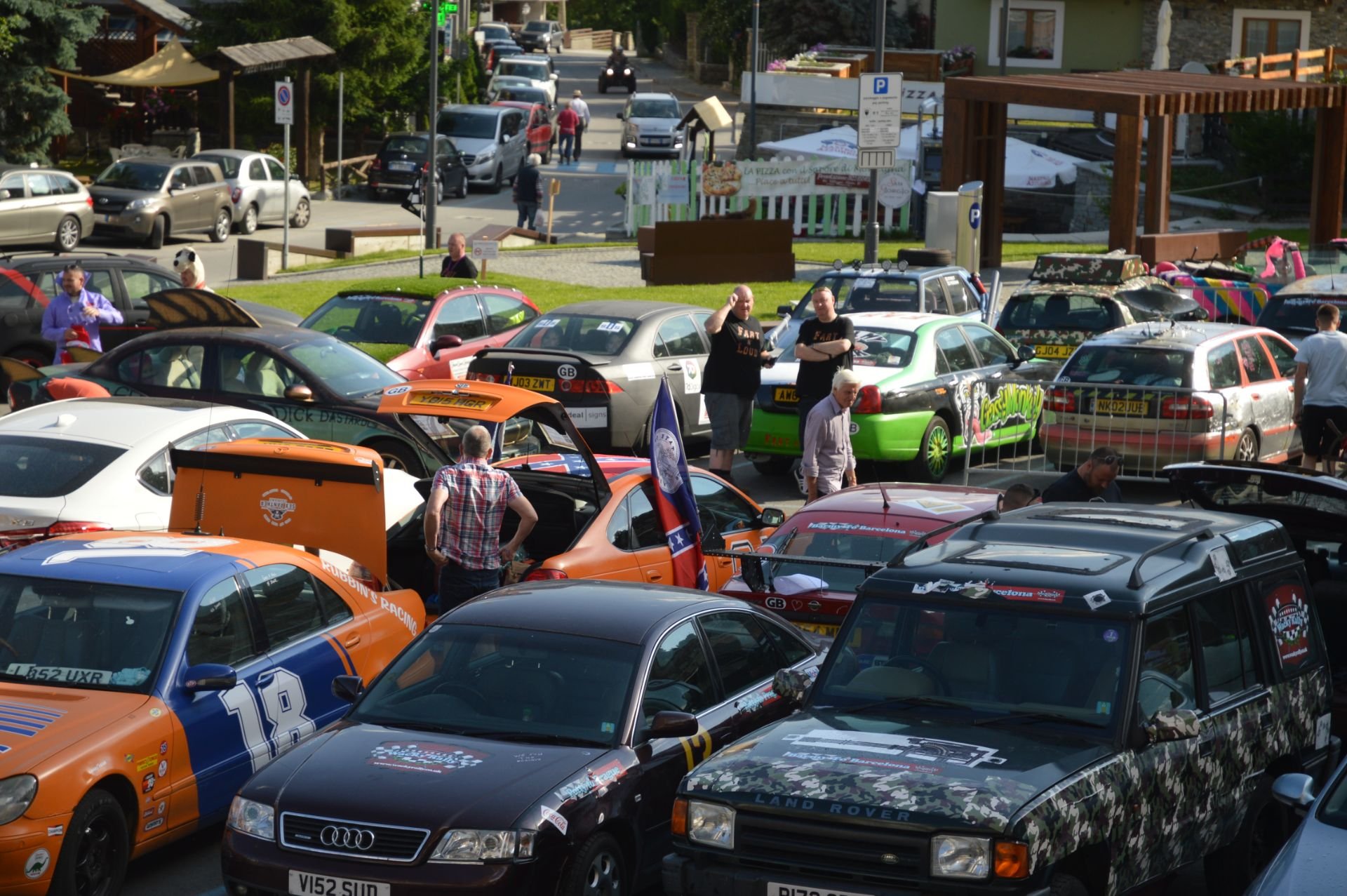 A gathering of cars and people in an outdoor parking area, with some cars open and people inspecting them, amidst a background of buildings and trees.