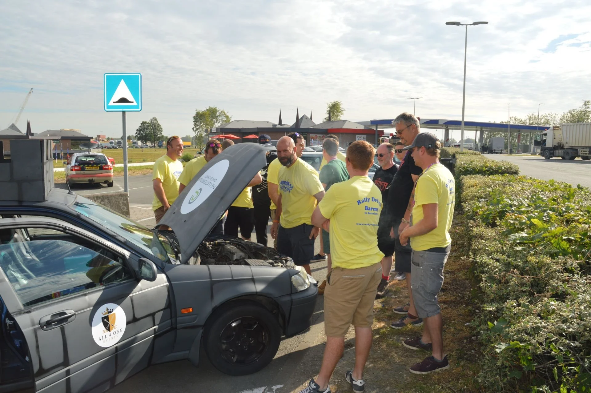 A group of people gathered around a grey car with an open hood in a parking lot, some wearing yellow shirts. The scene is set in daytime with a cloudy sky and a gas station in the background.