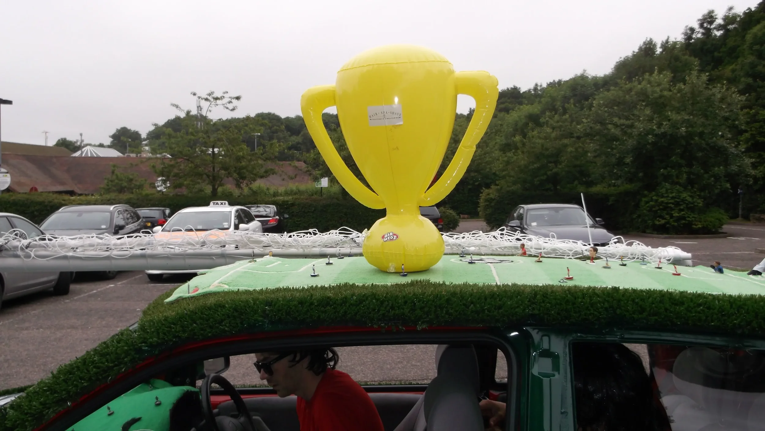 A large yellow trophy-shaped hot air balloon on top of a vehicle's roof, with a person sitting inside the vehicle, parked in a lot with other cars and trees in the background.