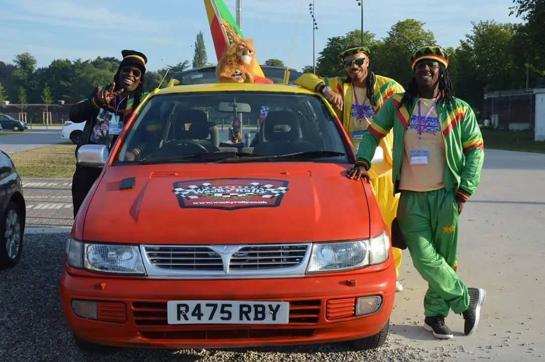 Four people in colorful Rastafarian-style clothing and hats posing around a red car with a lion mascot in the sunlit parking lot, with trees and a street in the background.