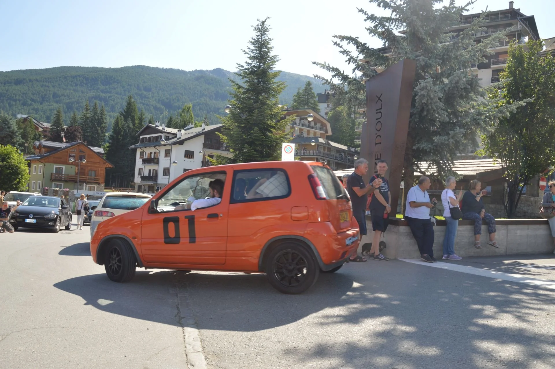 A small orange car with the number 01 on its side in a mountain town, with people sitting and standing nearby, and buildings and trees in the background.