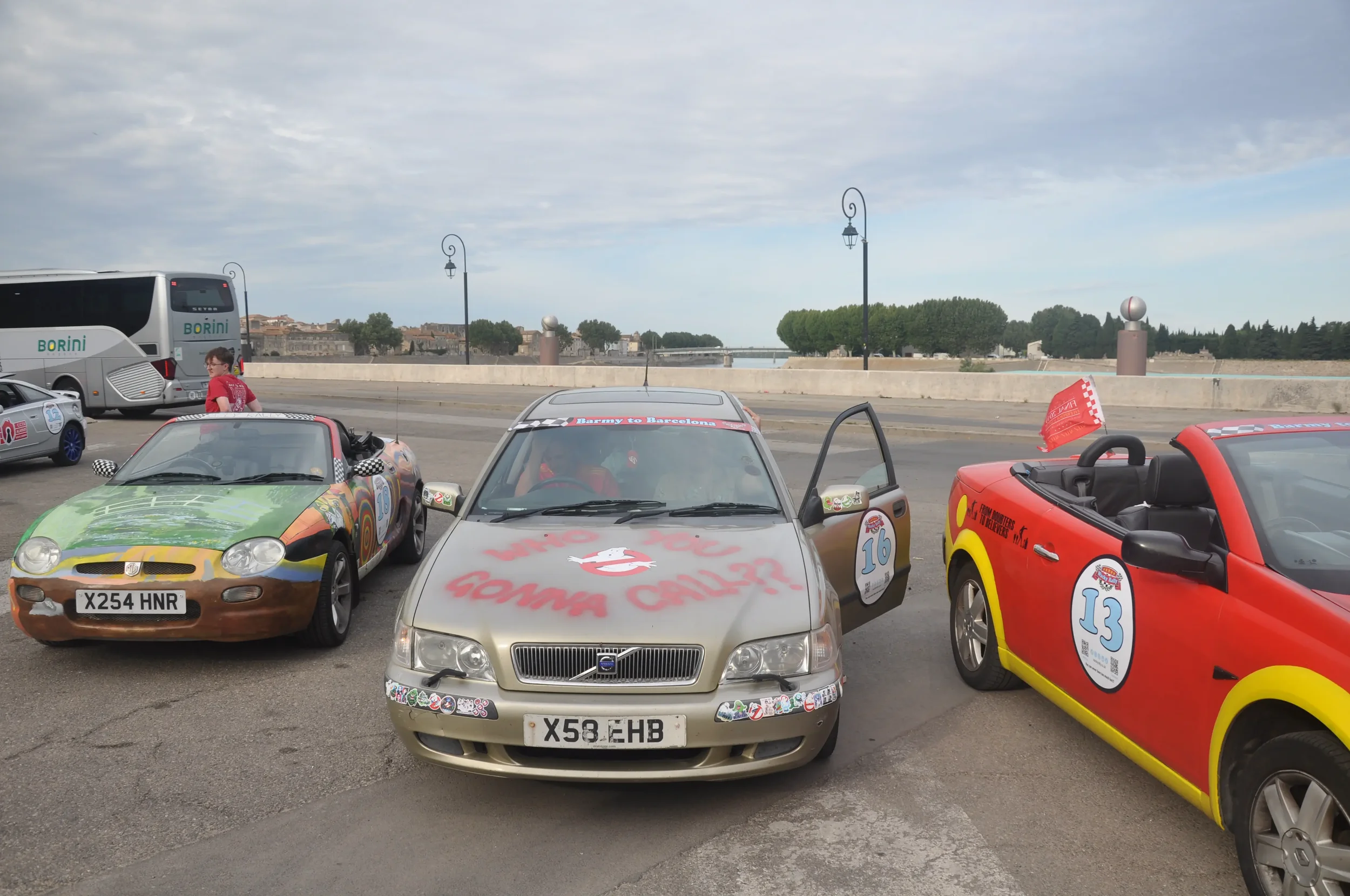 Several small toy or model cars parked outdoors on a paved surface, with a bus and a boy in the background. The cars have decorations and stickers, resembling racing cars with numbers and slogans.