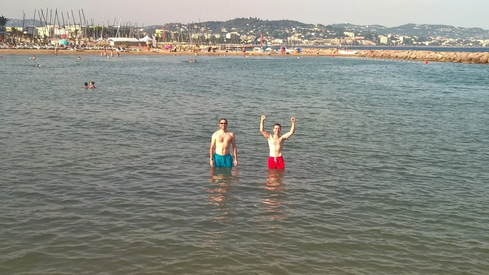Two men in swim trunks standing in the water at the beach, one with red trunks raising his arms triumphantly, with a sandy shoreline, people swimming, boats, and a hill with buildings in the background.