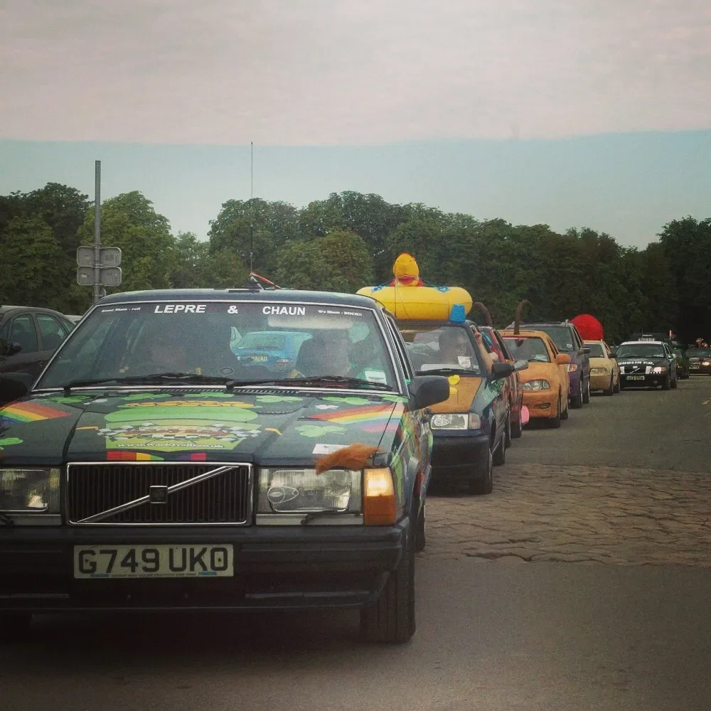 Line of colorful cars parked on a street, some with inflatable toys and accessories on top, and a green forest in the background.