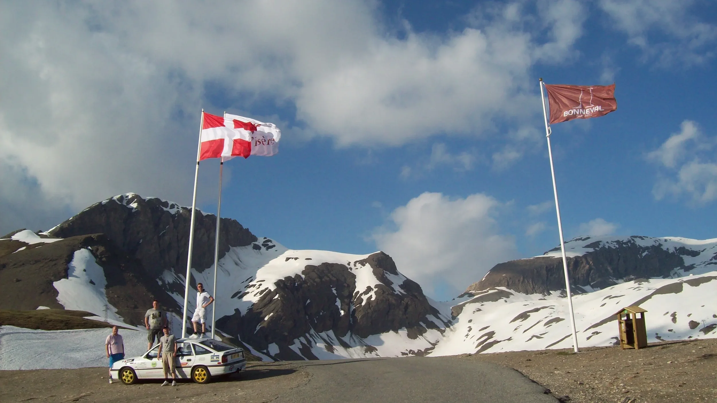 Four people standing near a small white car with yellow wheels, parked on a mountain road with snow patches. Three flags are flying in the background: one with a red and white cross, one with a red and white emblem, and another with a red background.