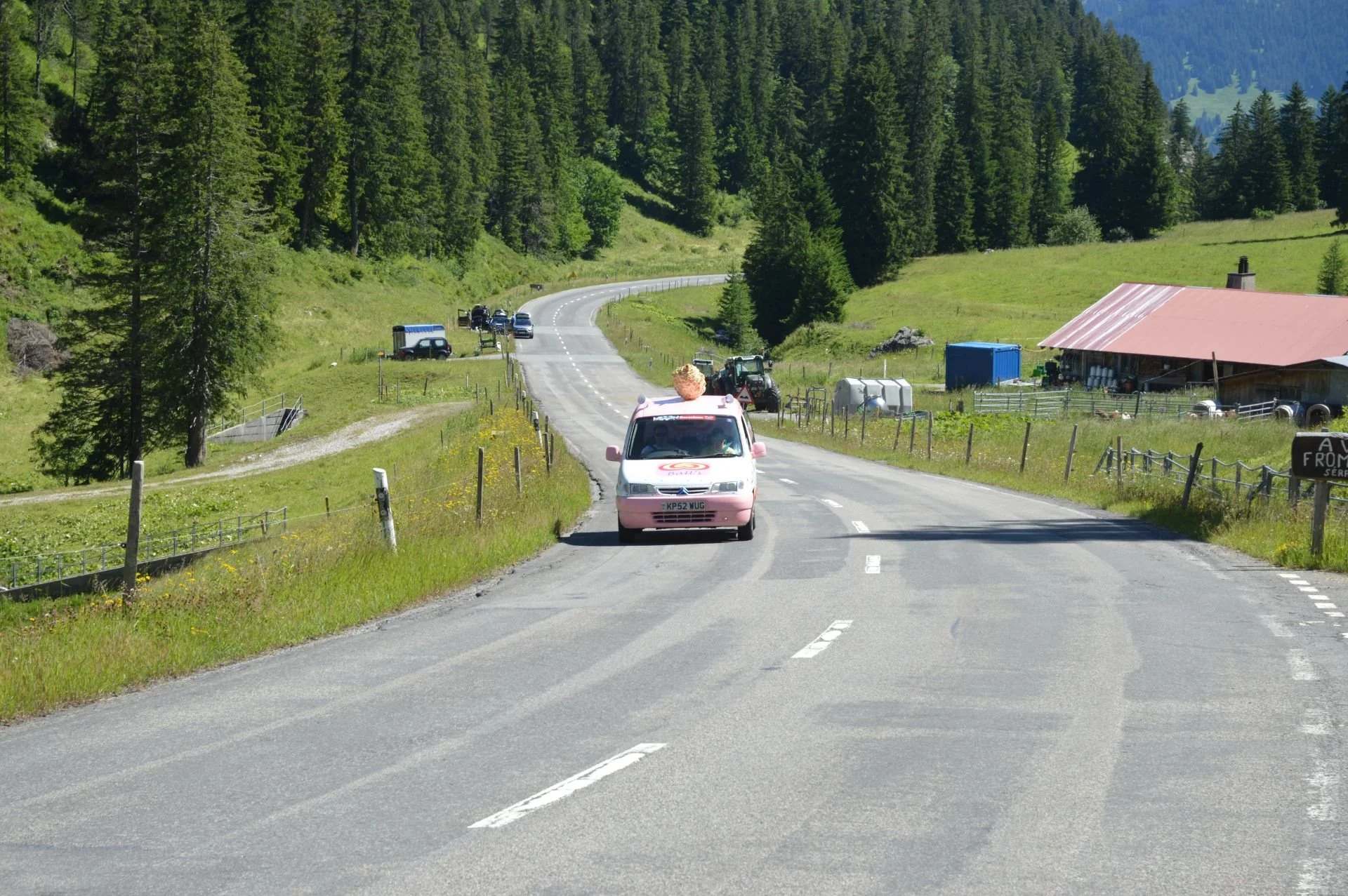 A rural mountain road with a pink food truck and a large ice cream cone on top of it, driving down the road, surrounded by green trees and hills, with farm buildings and tractors on the side.
