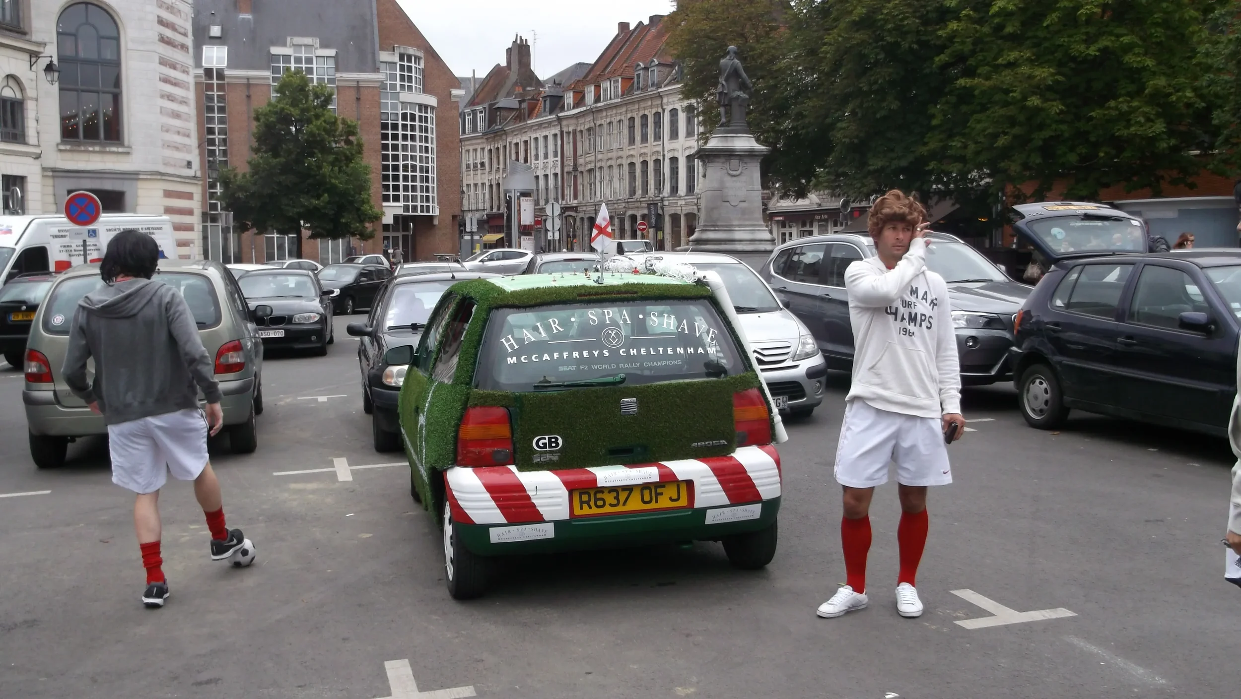 A small car decorated with artificial grass and red and white stripes, parked in a busy city parking lot with cars and buildings in the background. Two men in white sportswear and red socks stand nearby.