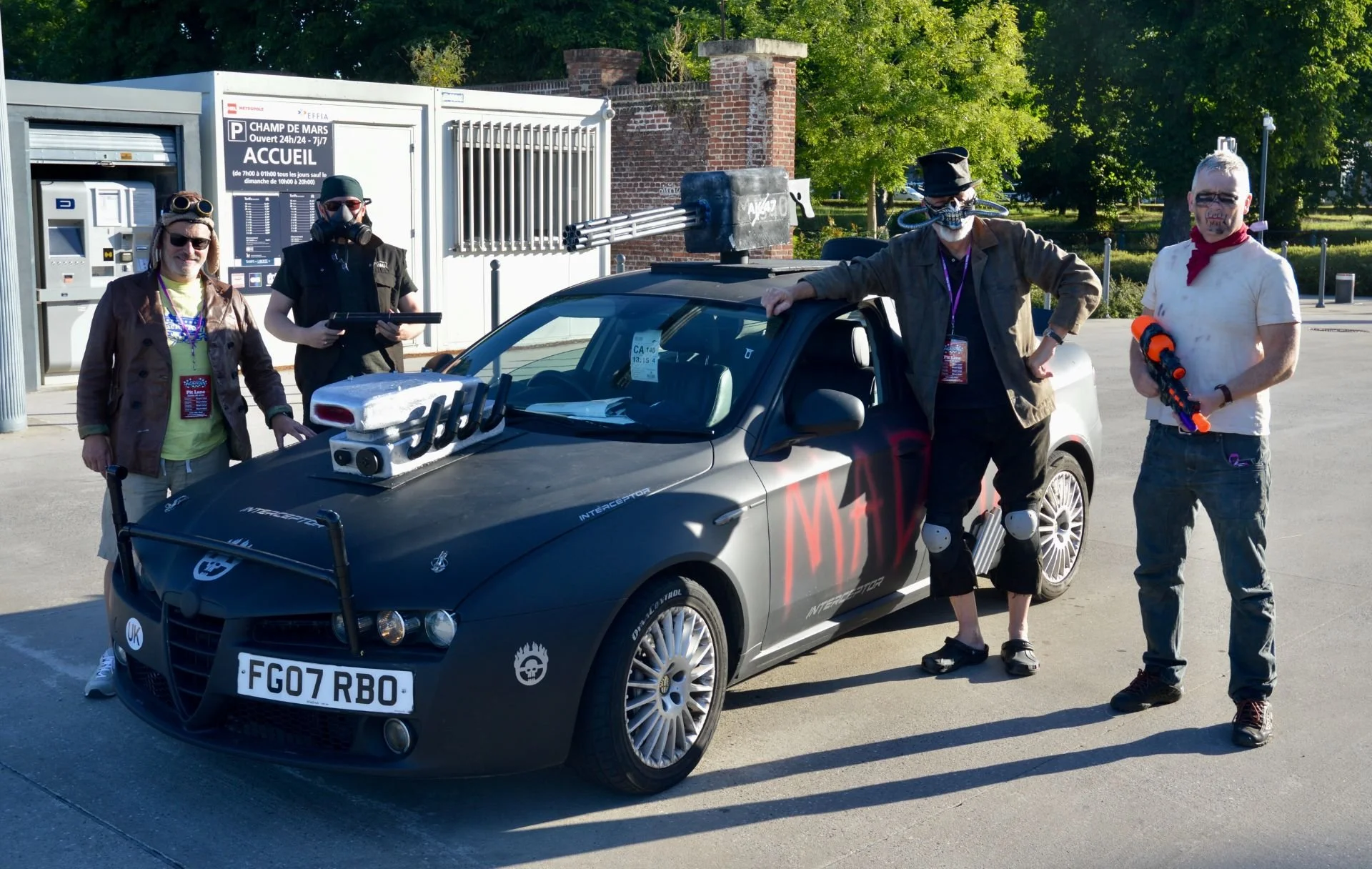 Group of five people in a parking lot with a custom modified car. The car is black with various props and decorations, including a large cannon on top, robotic arms, and the word 'MAD' painted on the side. Four of the people are wearing costumes or f