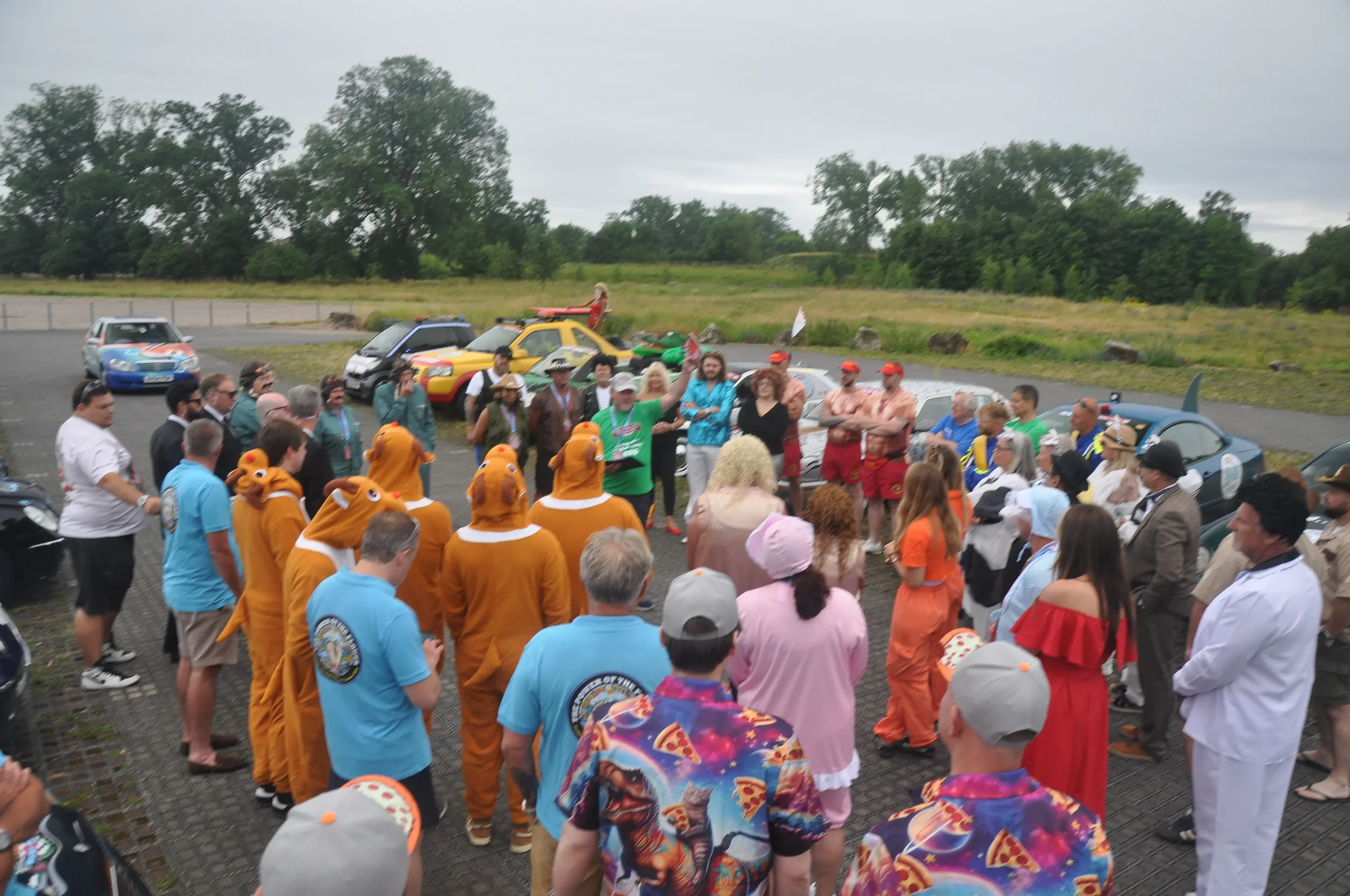 Group of people gathered outdoors in a parking lot, some in costumes including a large group dressed as cats, others in casual or themed clothing, with cars and a grassy field in the background.