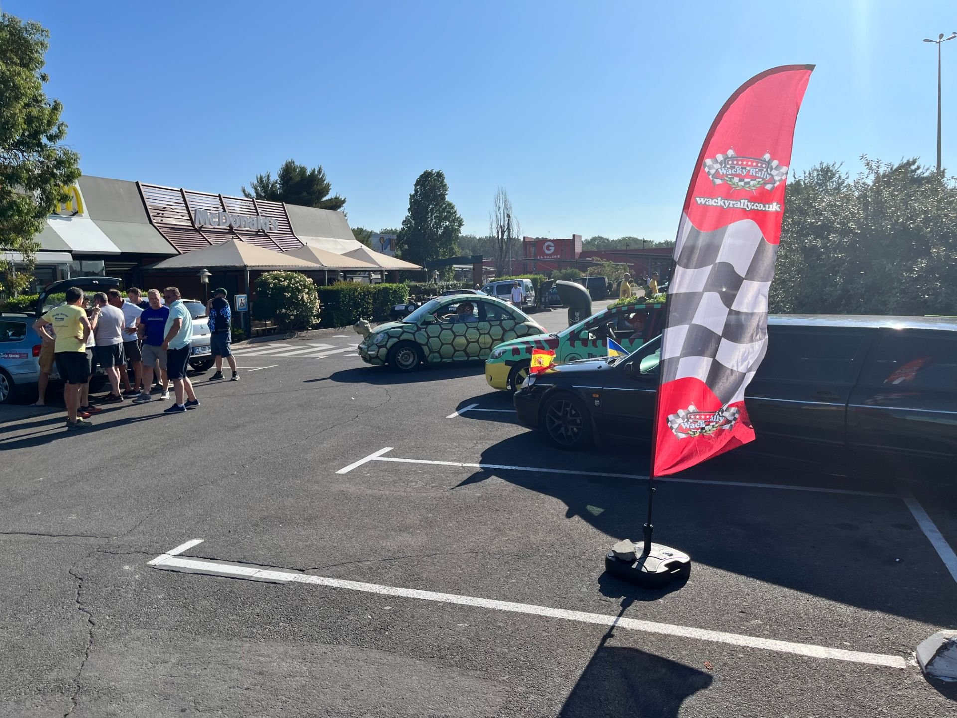 A parking lot scene with a group of people gathered near cars, a McDonald's restaurant in the background, and a Wacky Rally flag in the foreground.