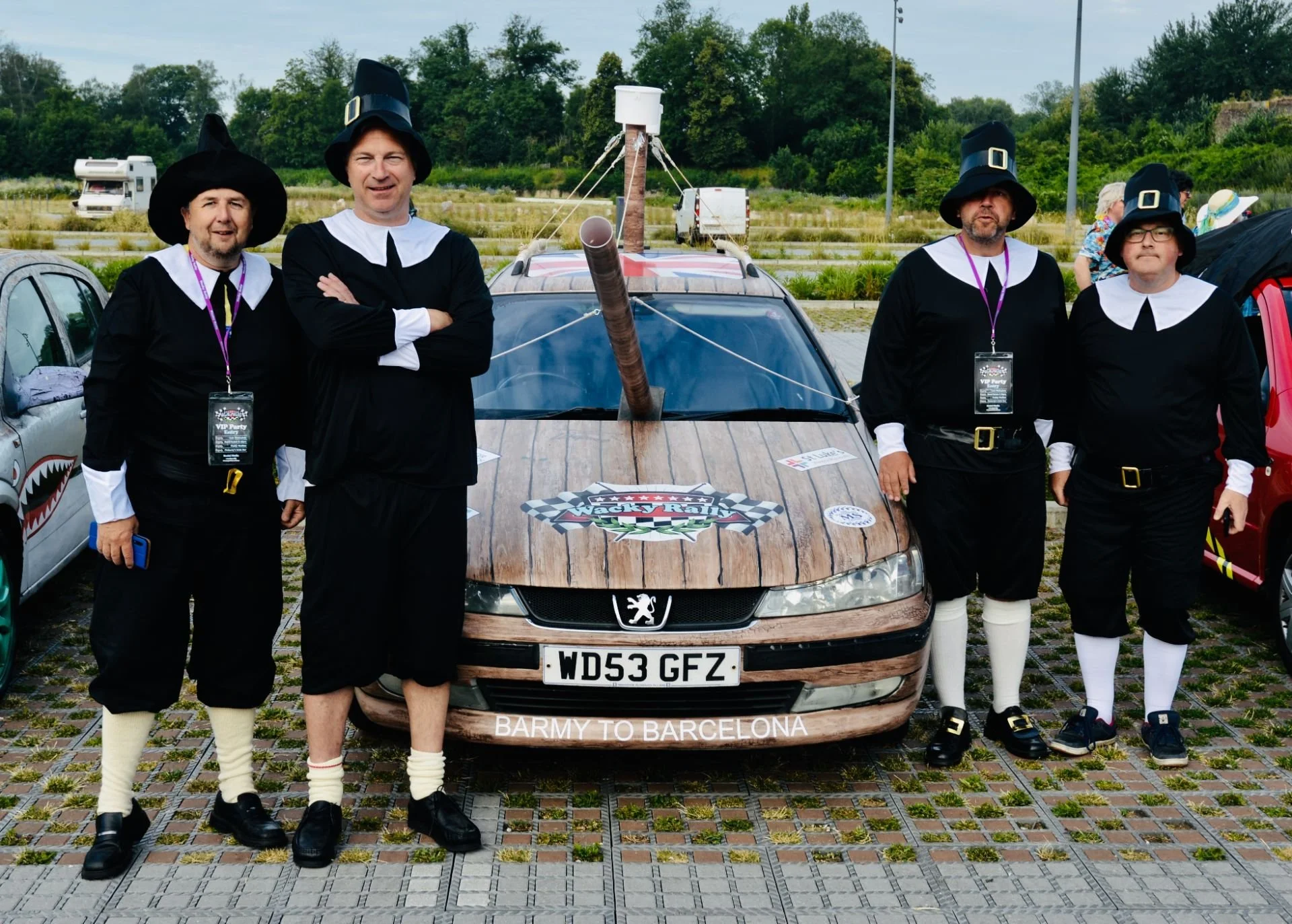 Four men dressed as witches in black robes, white collars, and witch hats standing in front of a car with wooden paneling. The car has a sign that reads "Jacky Kaly" and a bumper sticker that says "BARMY TO BARCELONA." They are outdoors in a parking 