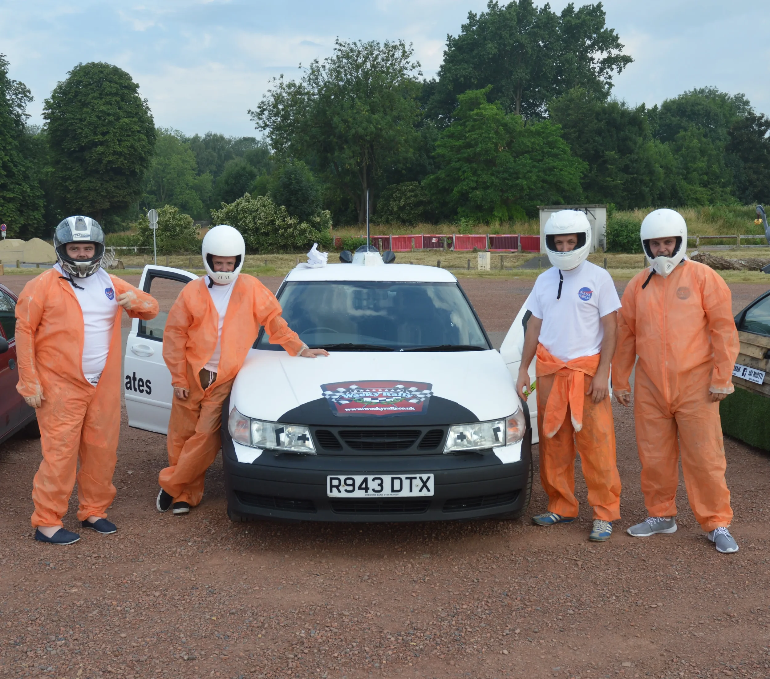 Five people in racing suits and helmets posing beside a satellite, race car in a gravel lot with trees in the background.