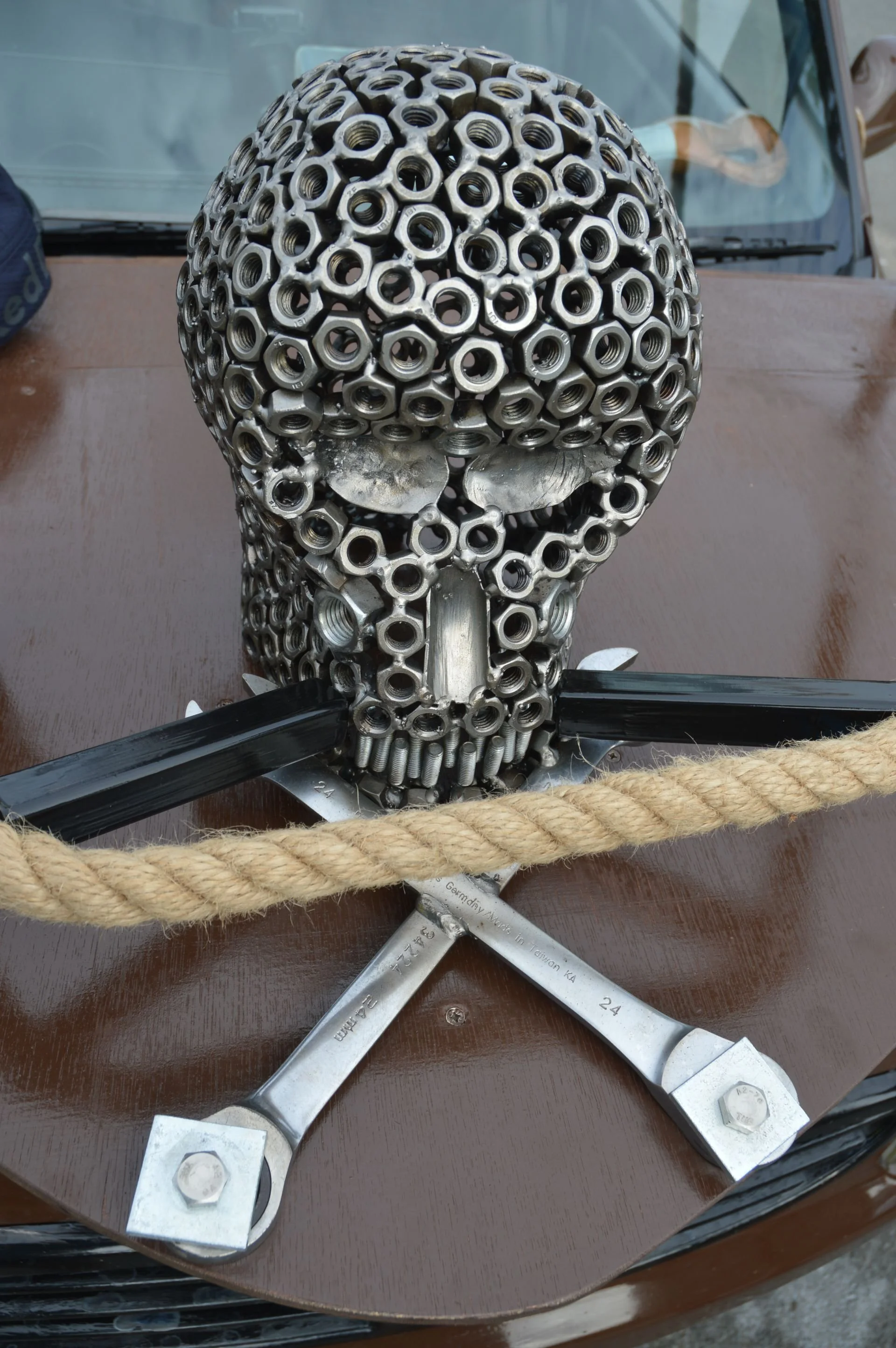 A skull sculpture made of metal nuts and bolts on a wooden table, with a wrench and a rope in the foreground.