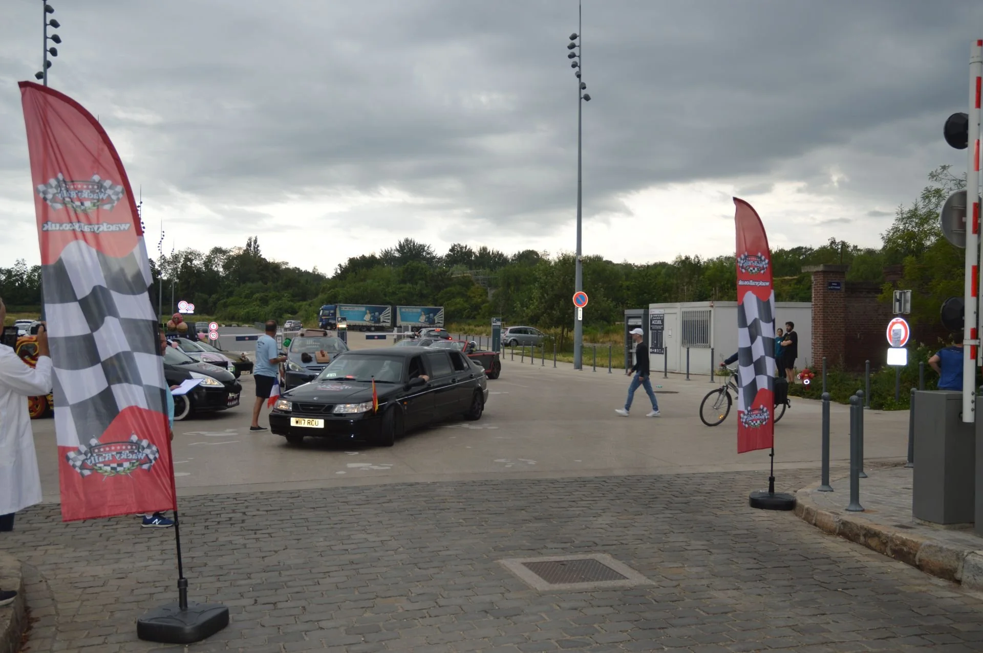 Several cars parked outdoors on a cloudy day, with people walking and standing around. Display flags with racing checkered patterns are visible, indicating a motorsport event.