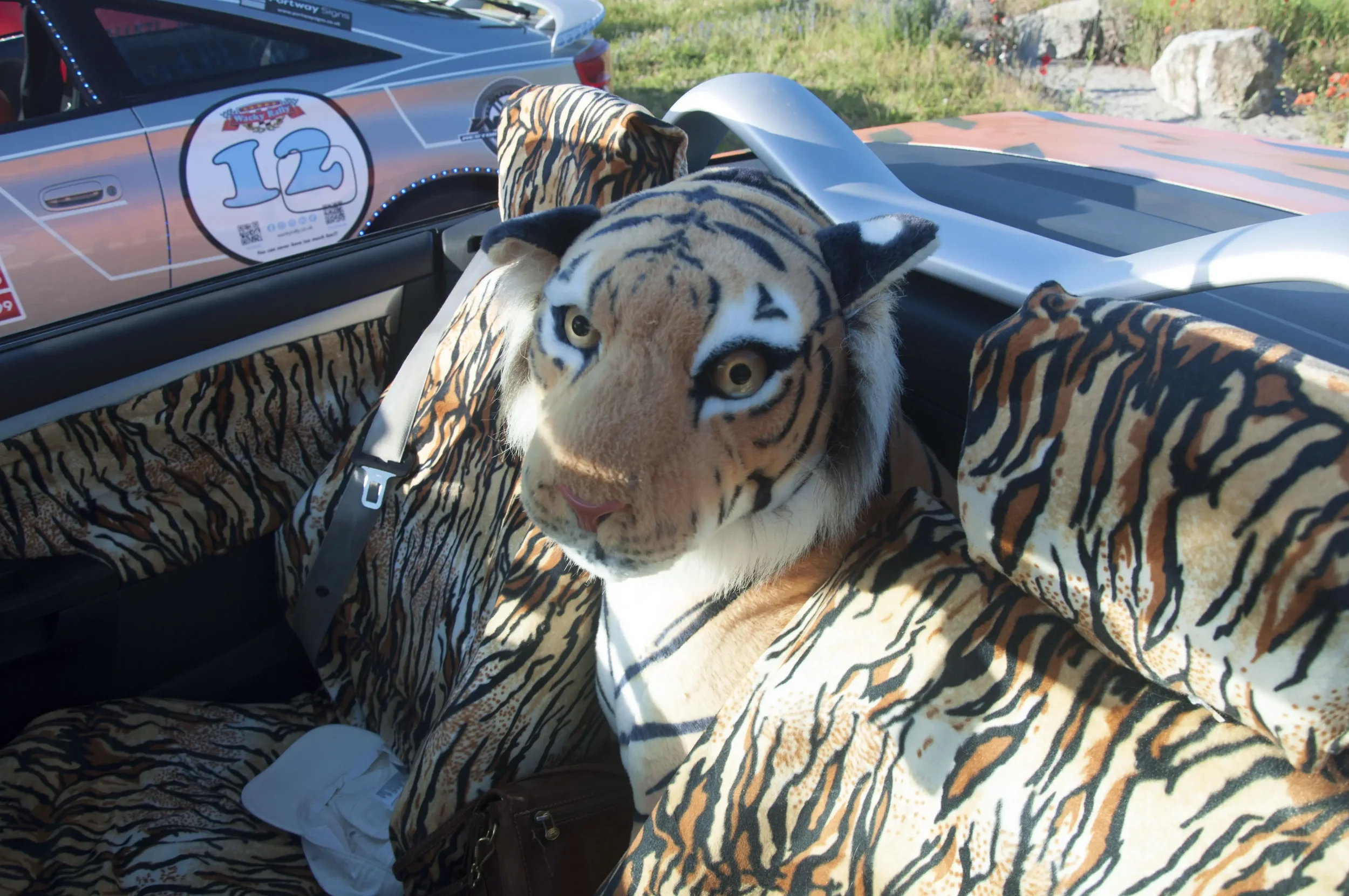 Person wearing a tiger costume seated inside a vehicle, with the costume's tiger print fabric covering the seat and part of the person's body. The costume includes a tiger headpiece with realistic features and markings, and the person is sitting in f