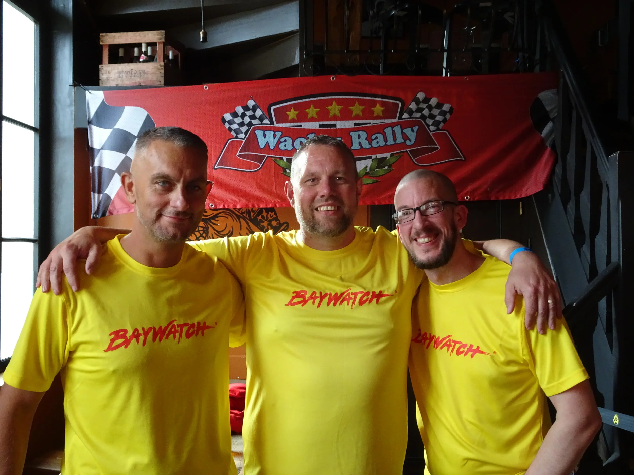 Three men wearing yellow 'Baywatch' shirts, smiling and standing arm-in-arm indoors with a red banner that says 'Wagon Rally' and checkered flags in the background.