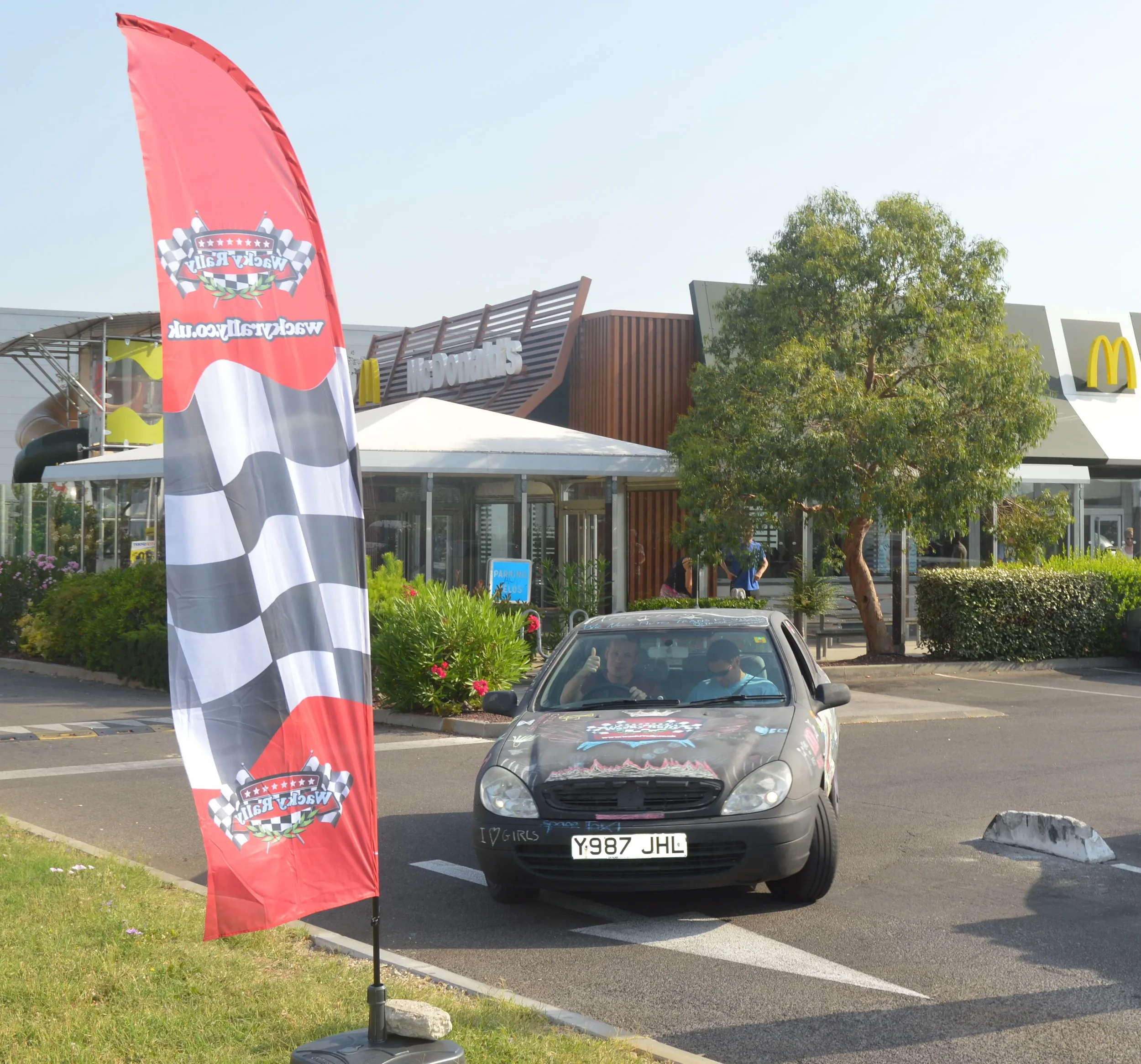 A car parked in a parking lot with a racing flag on the side, two people inside, and a McDonald's restaurant in the background.