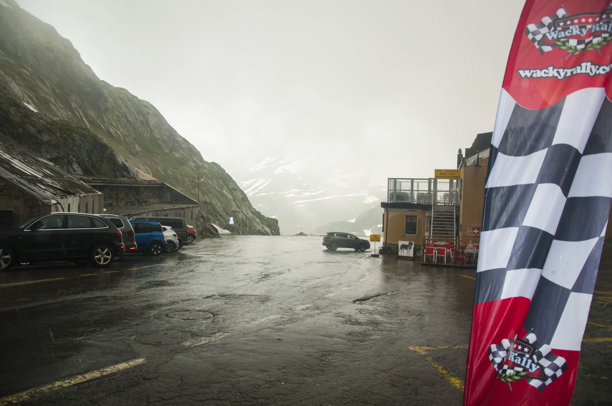 Rainy day at a mountain parking lot with wet pavement, mountains with snow patches in the background, several parked cars, a small building with outdoor seating, and a checkered flag banner with rally logo.