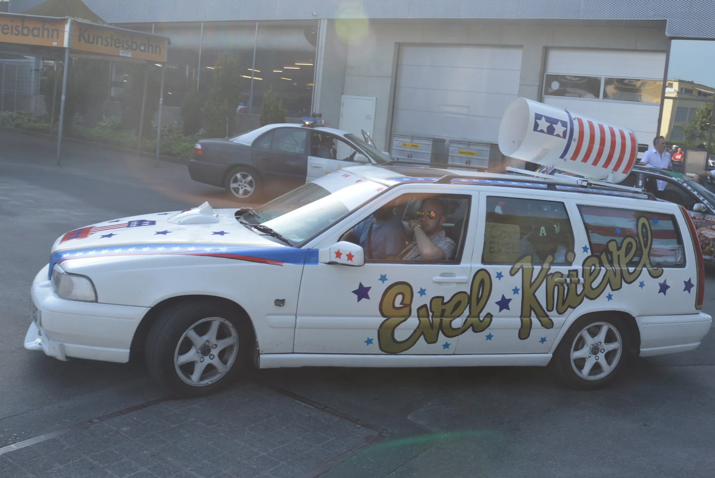 Decorated white car with 'Enel Knorvel' written on the side, featuring patriotic stars and stripes, and a large soda cup on the roof, parked outside a building near a police car, with two people inside wearing sunglasses and a sign in the back window