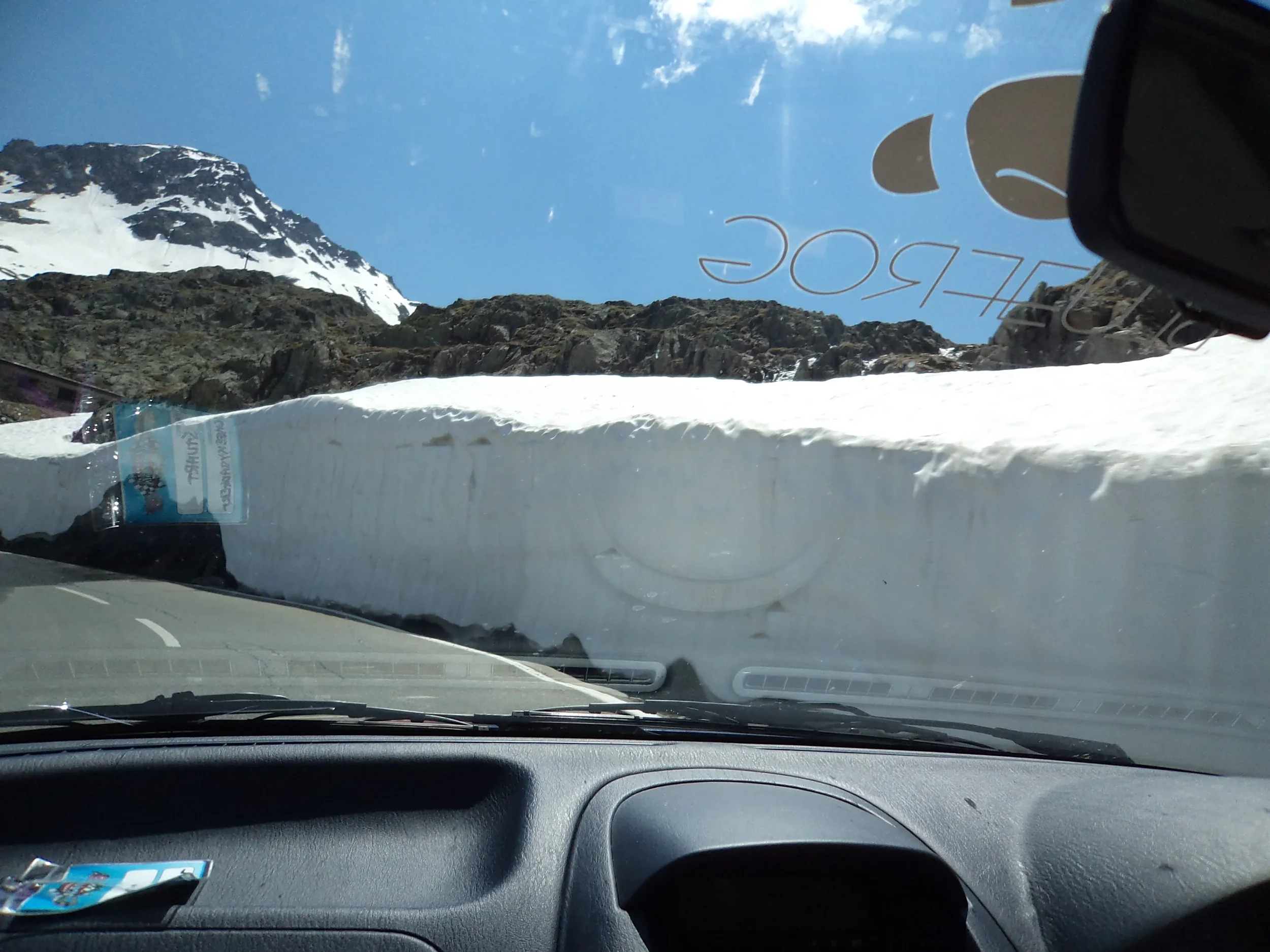 View from inside a vehicle showing a snowbank blocking part of the road, rocky mountains with snow, clear blue sky, and some reflections on the windshield.