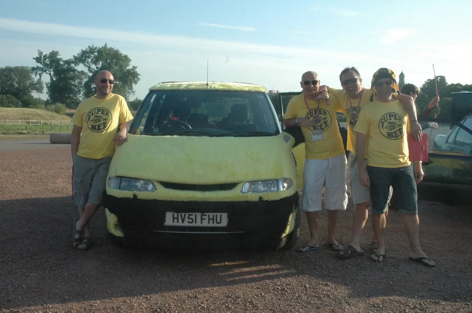 Group of five men wearing yellow t-shirts, standing outdoors next to a small, yellow car with a black bottom, in a parking lot with trees and open sky in the background.