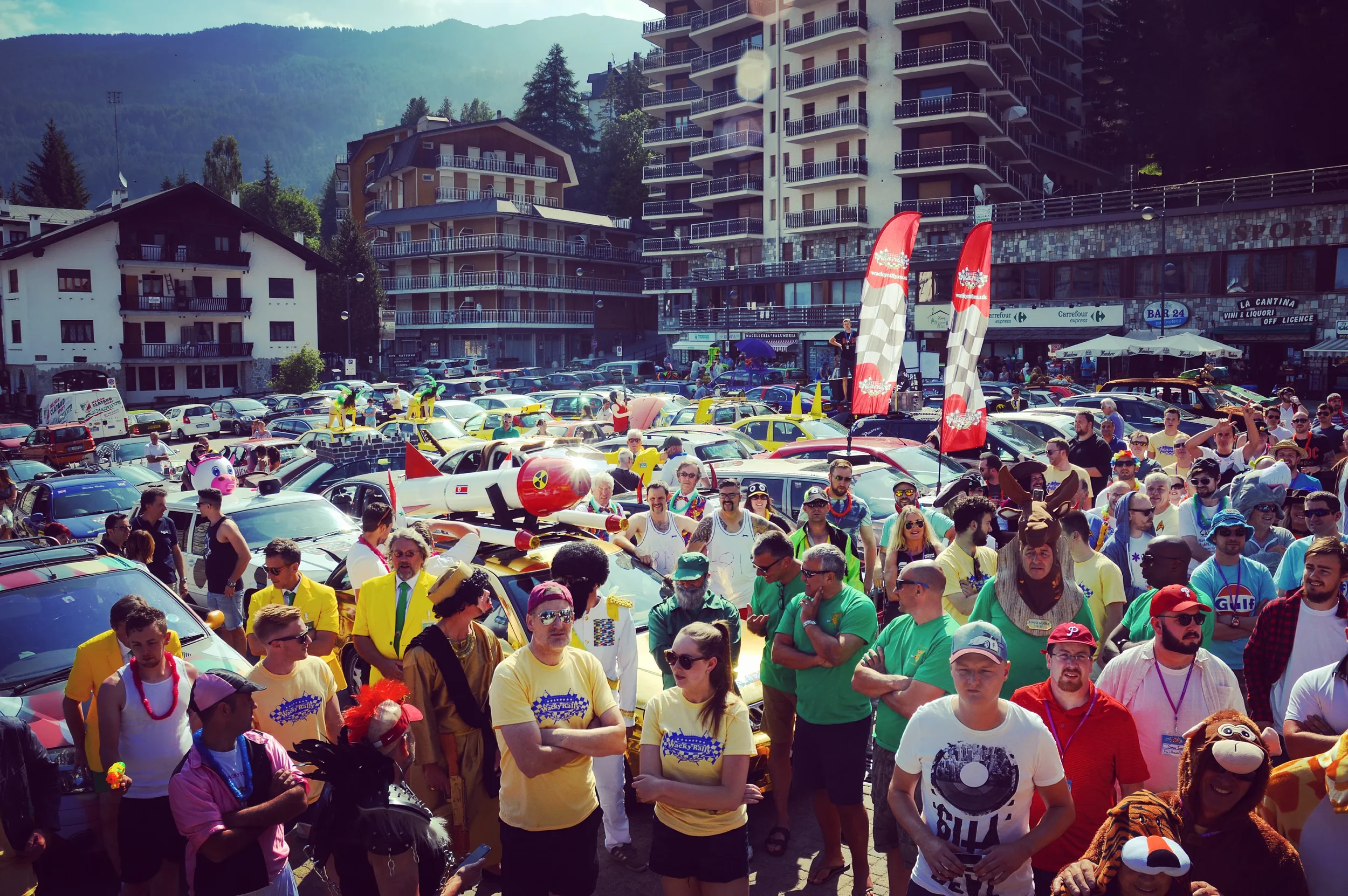 Crowd gathered at a car event in a town with buildings, mountains, and trees in the background. The scene includes people in colorful costumes, inflatable toys, flags, and decorated cars.