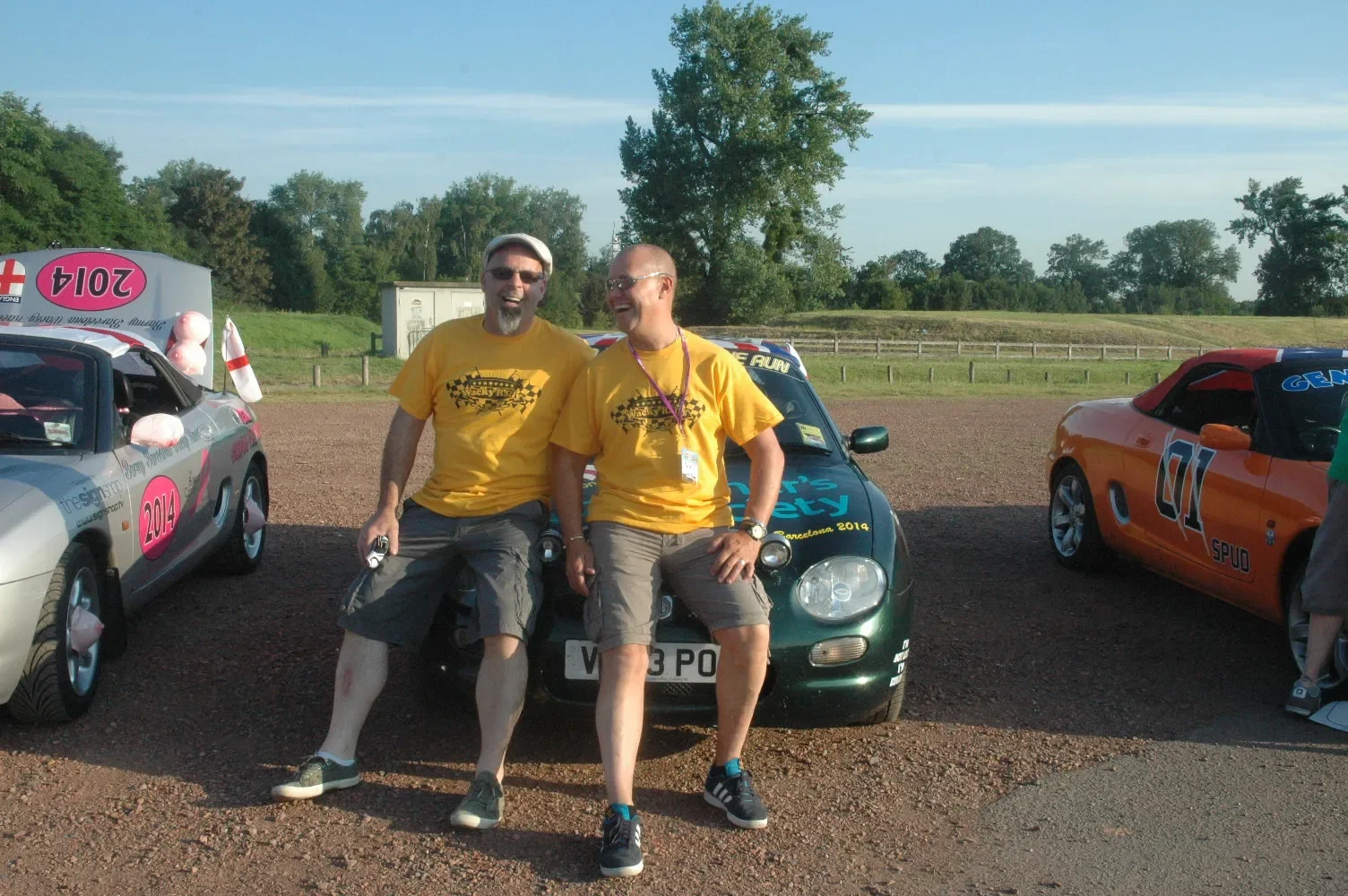 Two smiling men wearing yellow shirts with checkered flags and text, sitting on a green car, with two other decorated cars on either side, in an outdoor setting with greenery and clear sky.