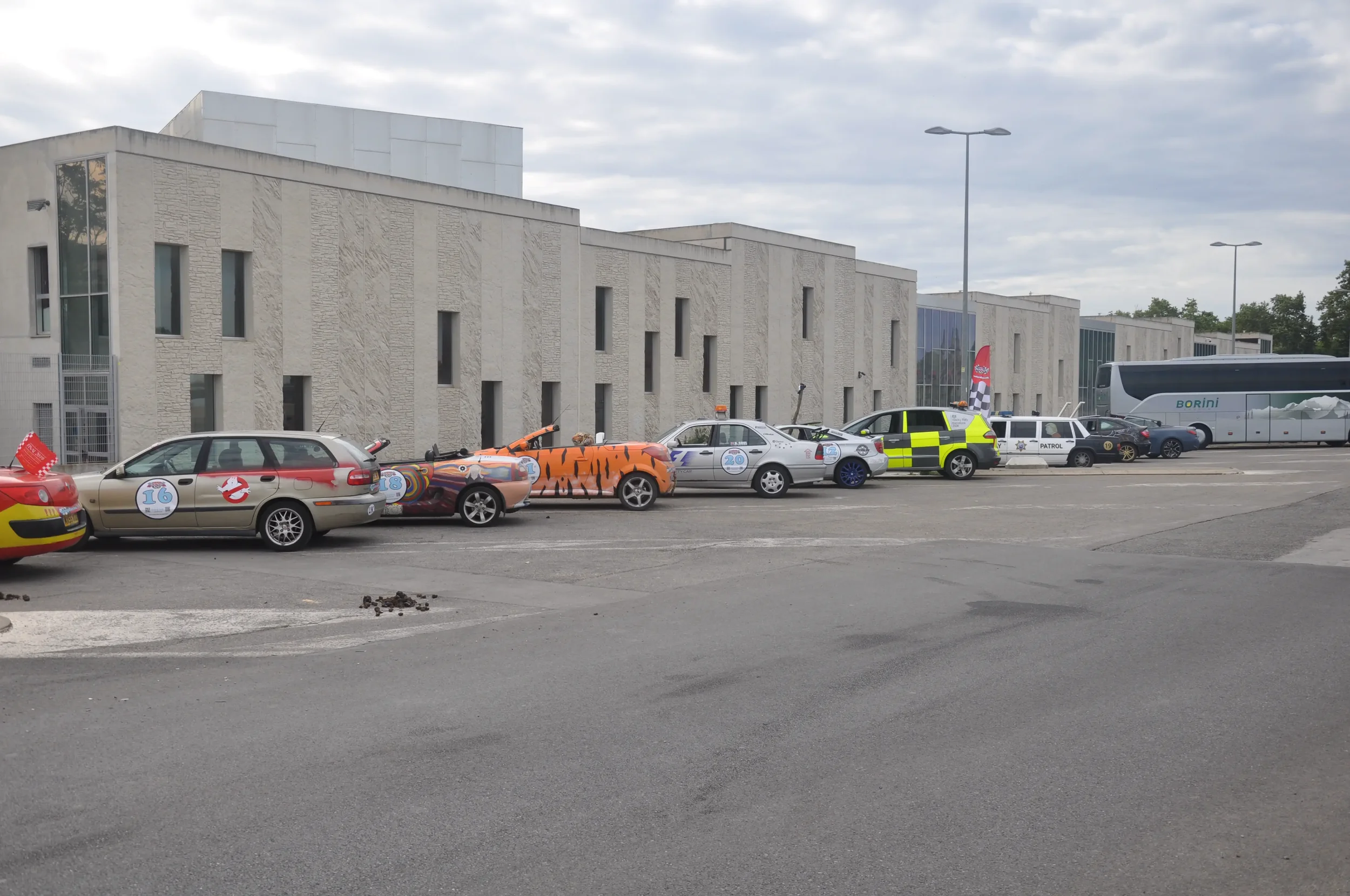 Line of parking spaces with various decorated cars, a police vehicle, a bus, and a modern building in the background under cloudy sky.