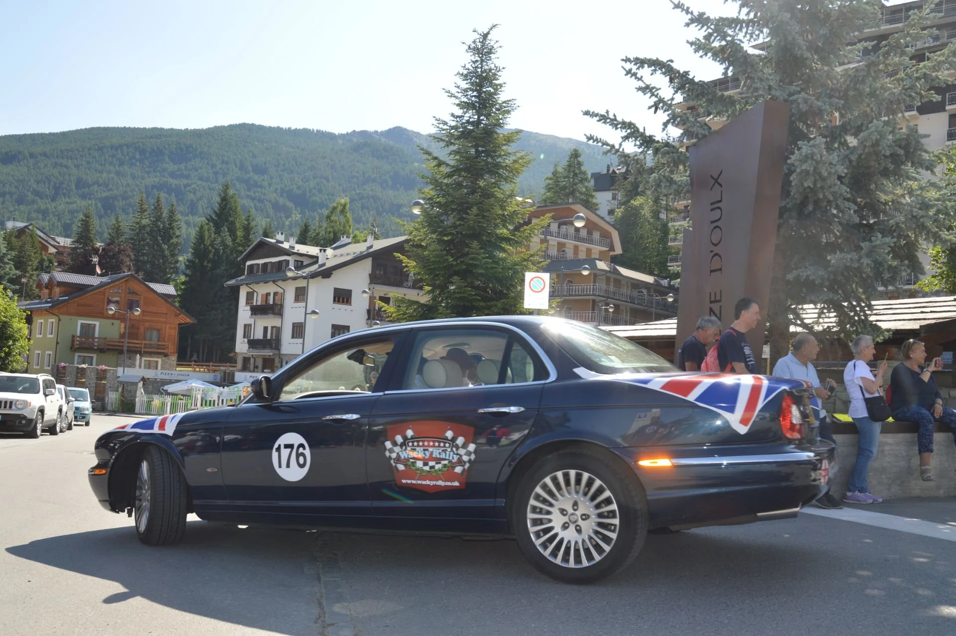 A dark blue luxury car with a racing number 176 on the door parked on a street in a mountain village, with groups of people sitting and standing nearby and a sign that reads 'LA DOUX' in the background, under clear skies with green mountains and resi