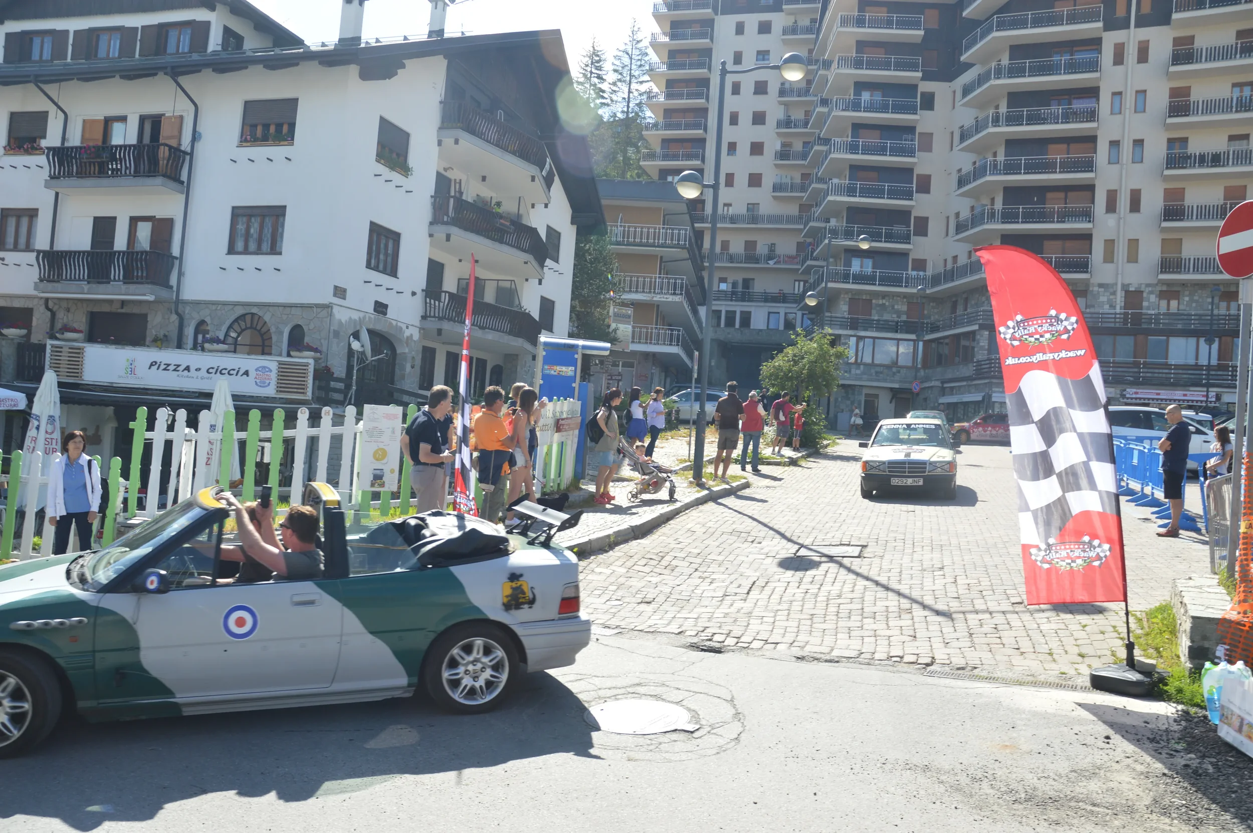 A street scene with a vintage car, quad racing flag, and people gathered around, in front of residential buildings.