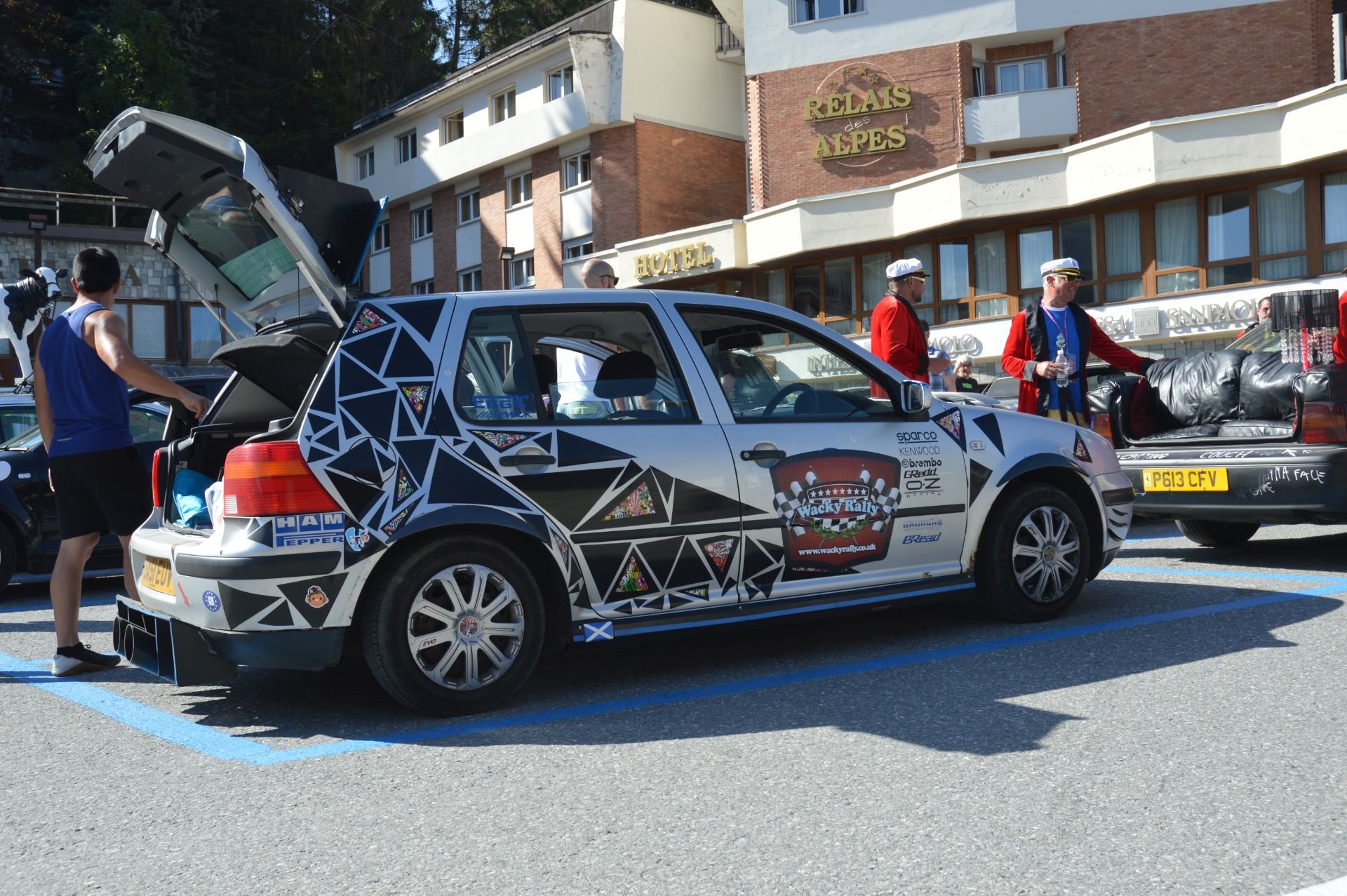 A decorated rally car parked in a lot with people around, some in racing attire, and buildings including a hotel behind it.