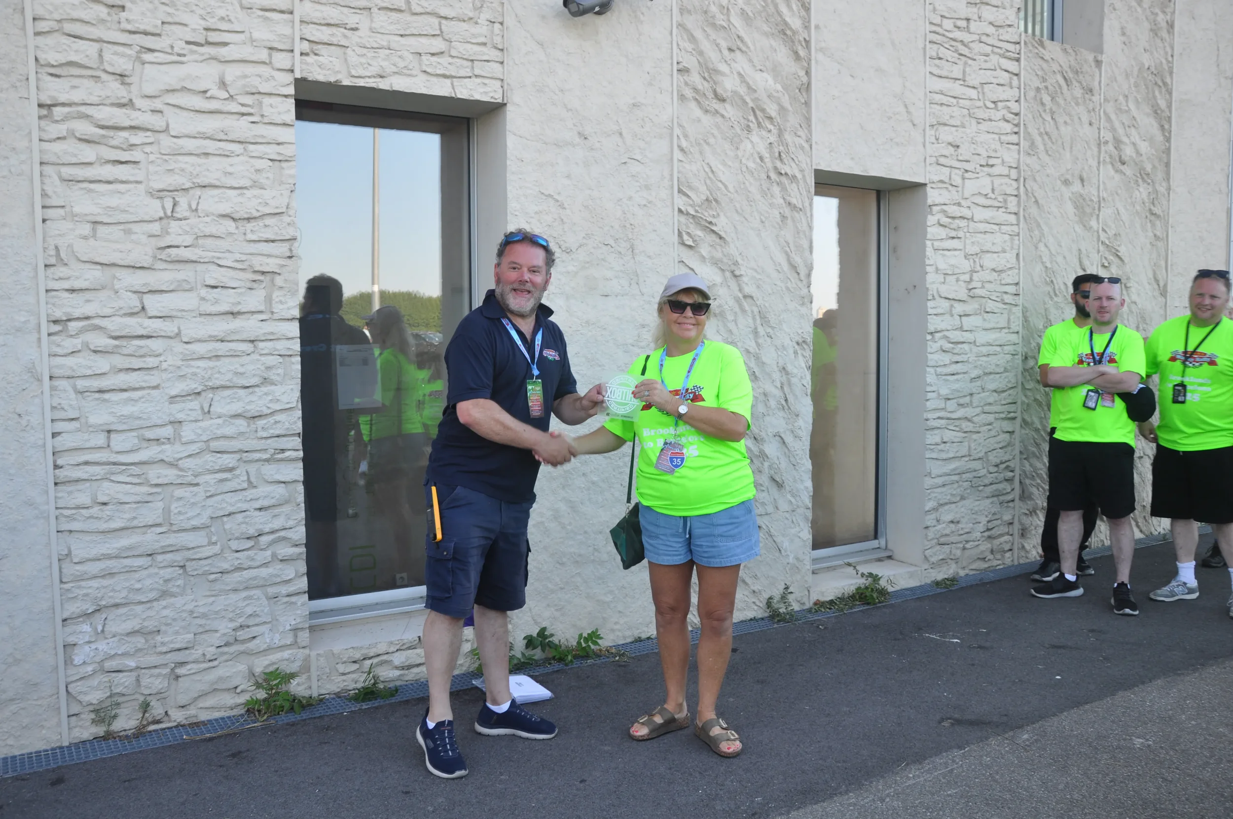 A man and woman shaking hands outside a building, with other people in bright yellow shirts standing in the background.