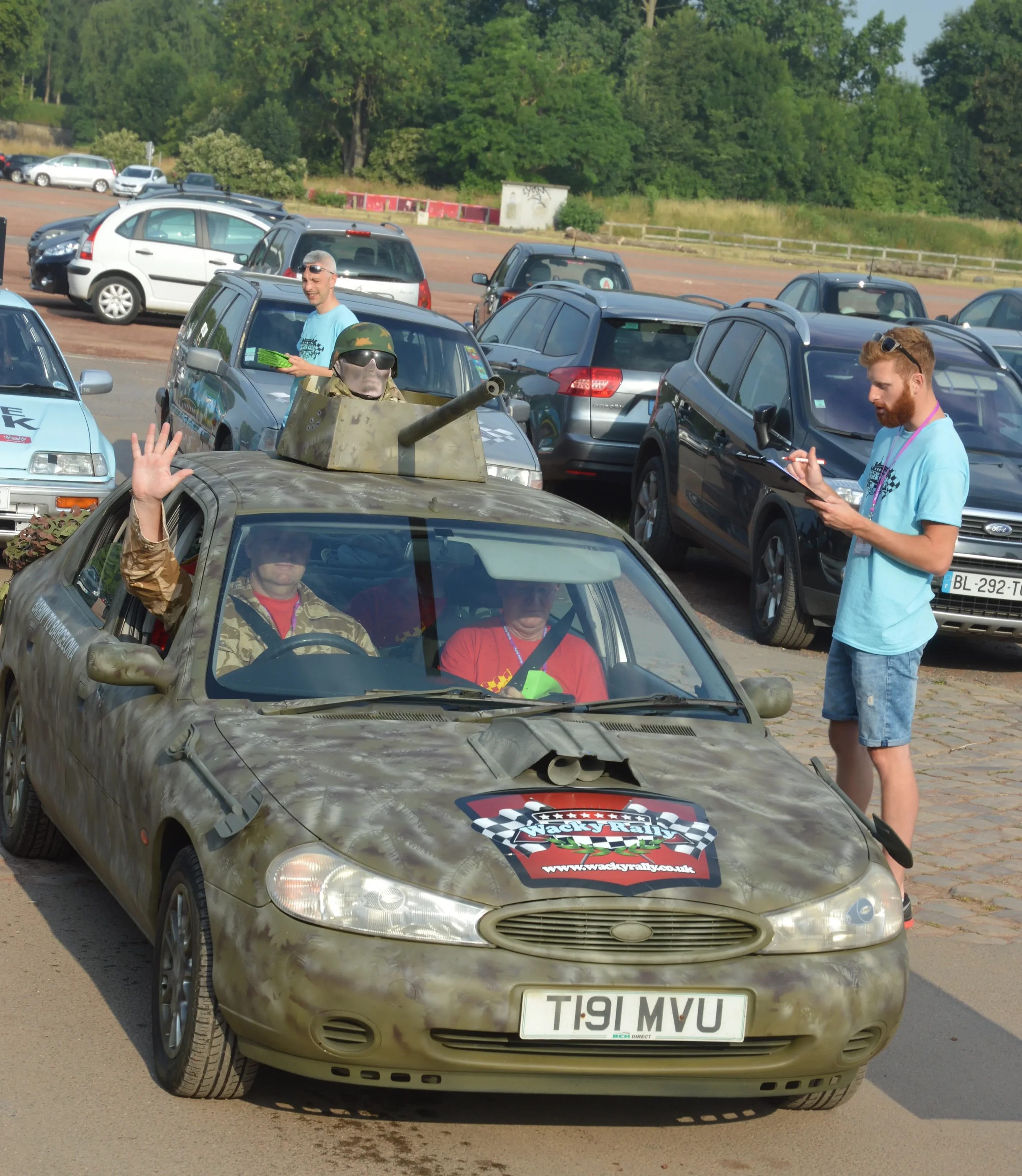 A car with a camouflage pattern and a 'Wacky Rally' decal on the hood, with two people inside; one waving and the other driving. Two men in blue shirts are standing nearby, one taking notes and the other holding a clipboard. There are several parked 