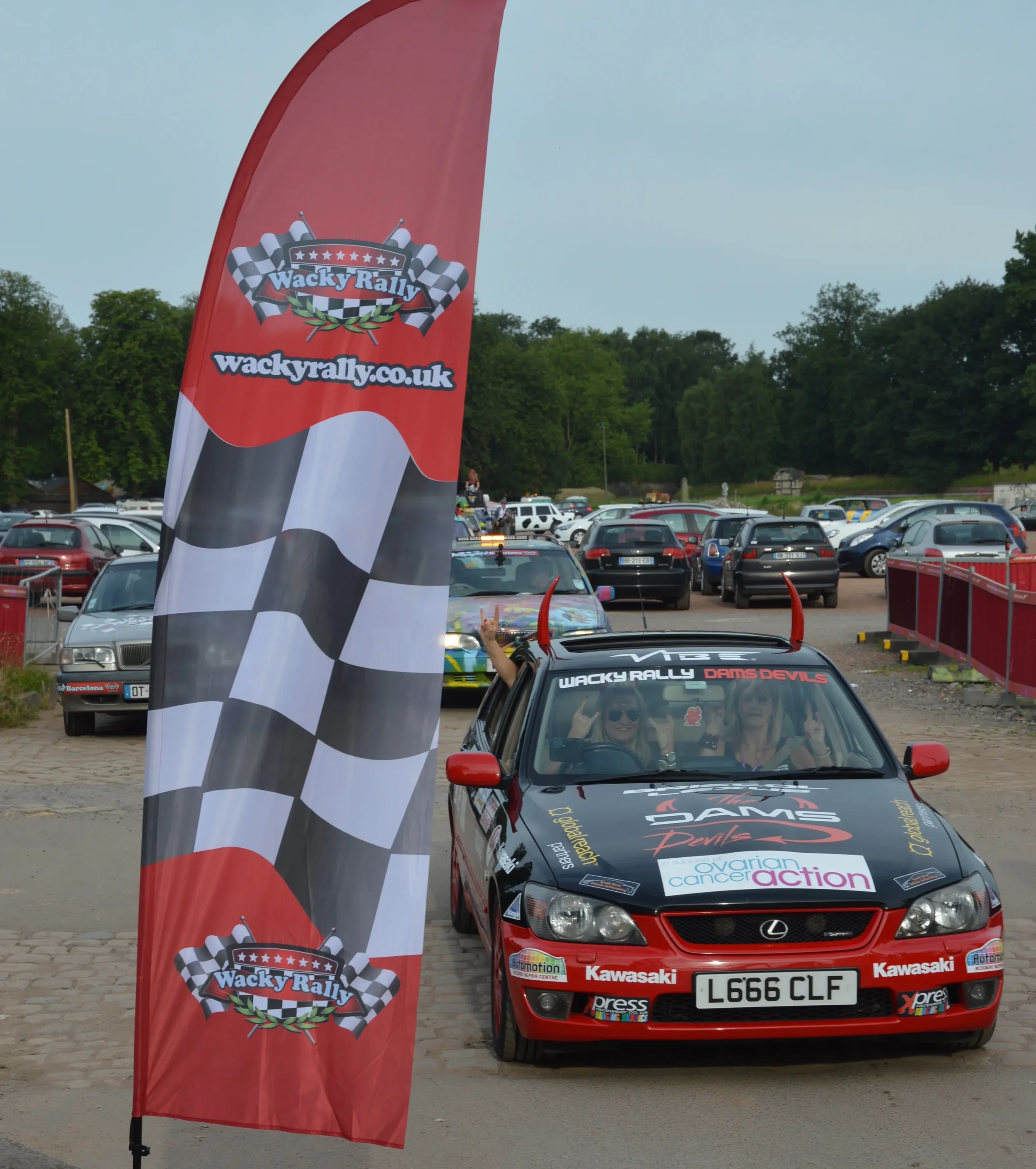 A red car decorated with various stickers and slogans, with two women inside making victory signs and wearing sunglasses, parked near a Wacky Rally banner with checkered flags, at a busy parking lot.