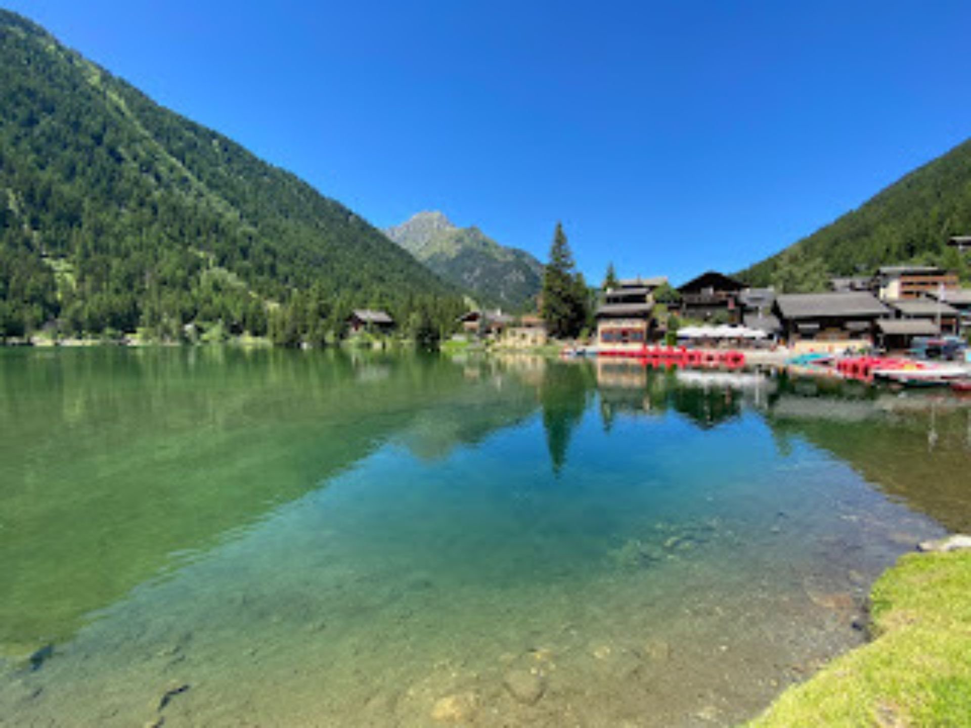 A lakeside scene with a small village at the base of green mountains under a clear blue sky, with calm water reflecting the houses and trees.