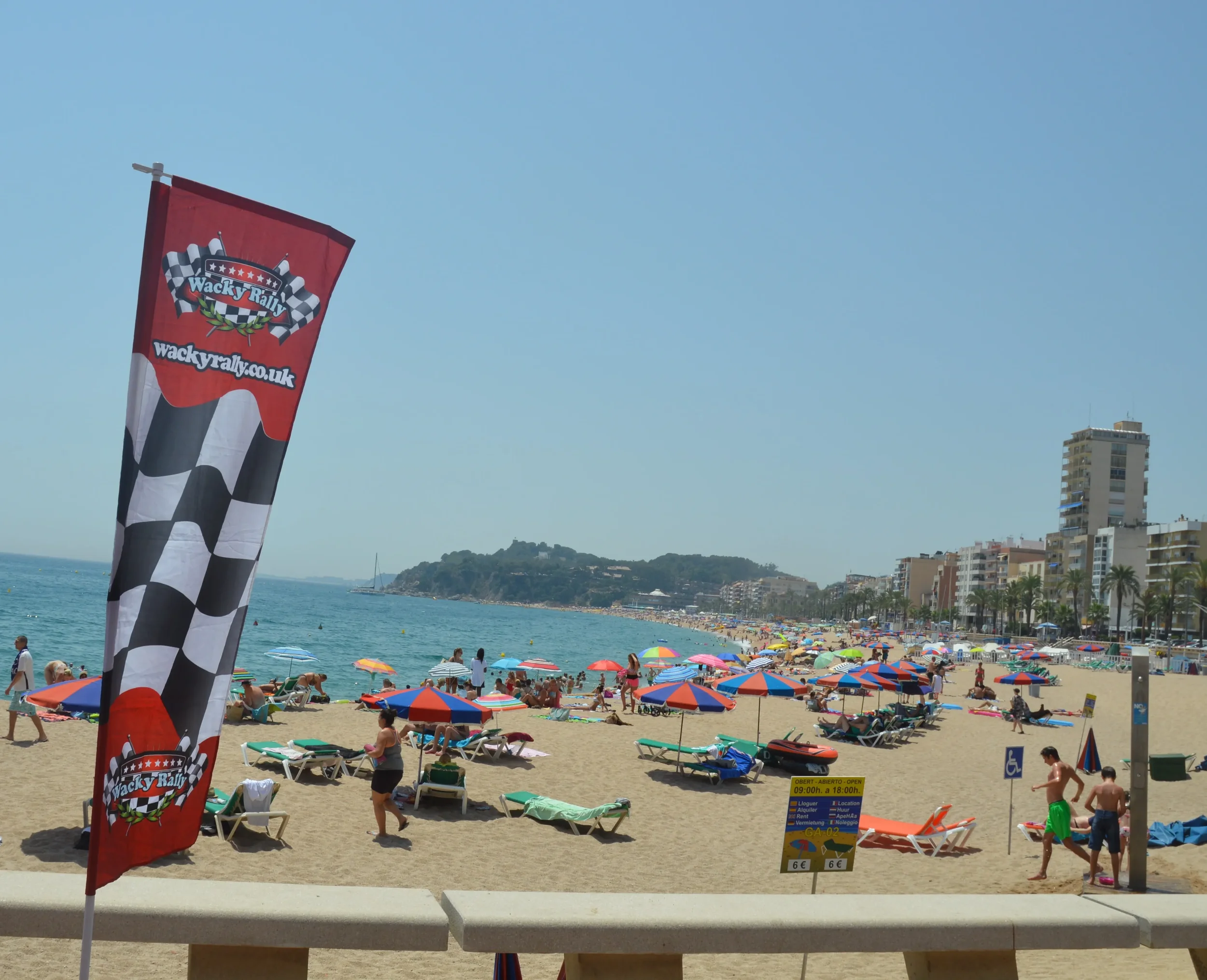 Beach scene with colorful umbrellas, beach chairs, and people walking along the shore under a clear blue sky. There are high-rise buildings and a hill in the background.