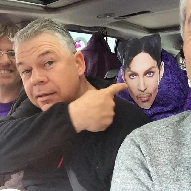 A man on a bus pointing to a cardboard cutout of a person with dark hair, purple outfit, and makeup, with other people visible in the background.