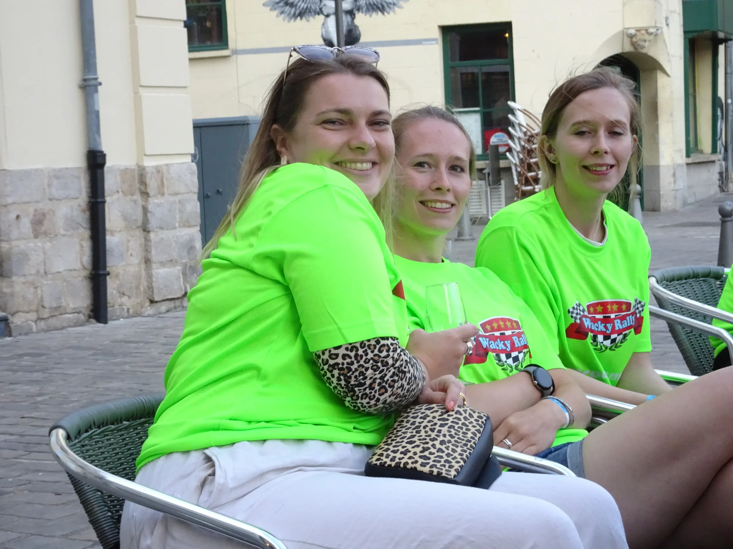 Three young women sitting outdoors on chairs, wearing bright green t-shirts with a racing-themed logo that reads "Wacky Rally". They are smiling, with one holding a wine glass. The background has a brick building with windows and some outdoor furnitu