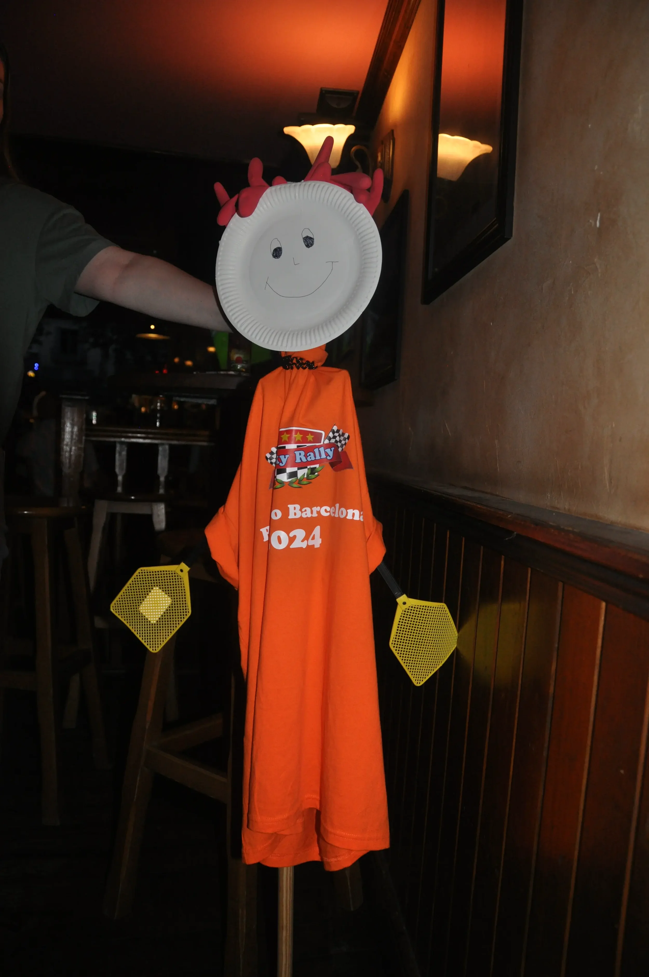 A paper plate with a smiling face drawn on it, attached to a scarecrow-like figure draped in an orange shirt, with yellow mesh fins on each side, inside a dimly lit room.